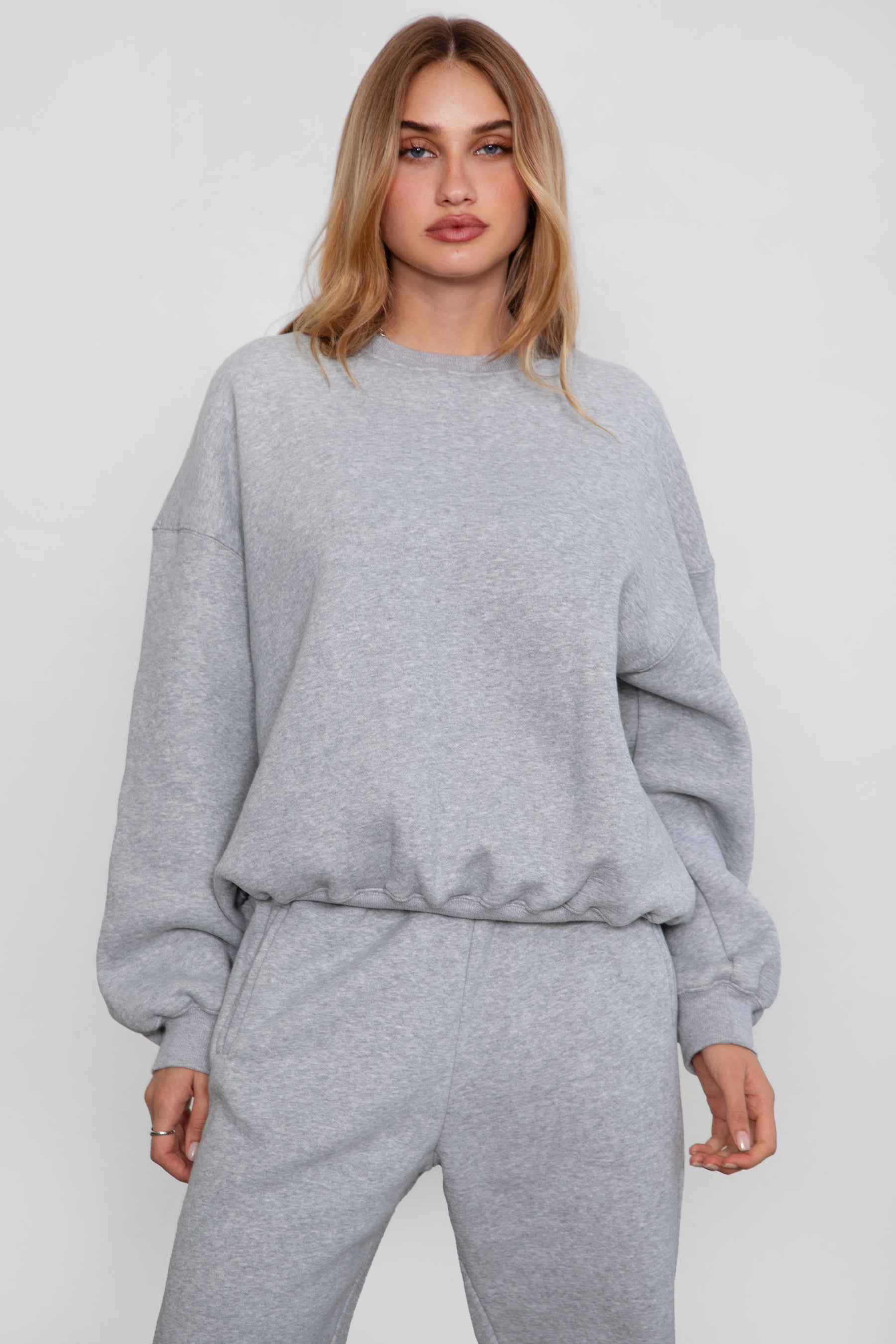 Woman wearing the TIC TOC TH28243 VARSITY heather grey fleece crew neck pullover sweatshirt featuring dropped shoulders, ribbed trim, and plush fleece fabric with the matching bottoms