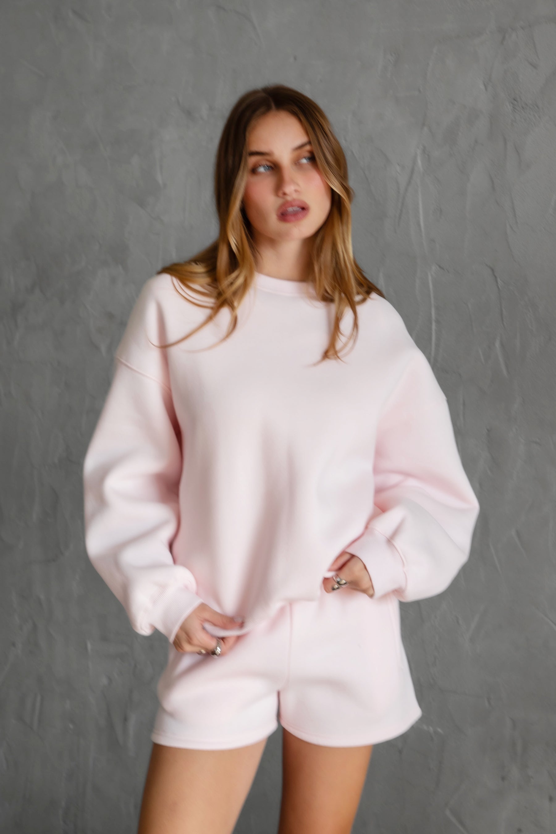 Woman wearing the TIC TOC TH28243 VARSITY pale pink fleece crew neck pullover sweatshirt featuring dropped shoulders, ribbed trim, and plush fleece fabric with the matching bottoms