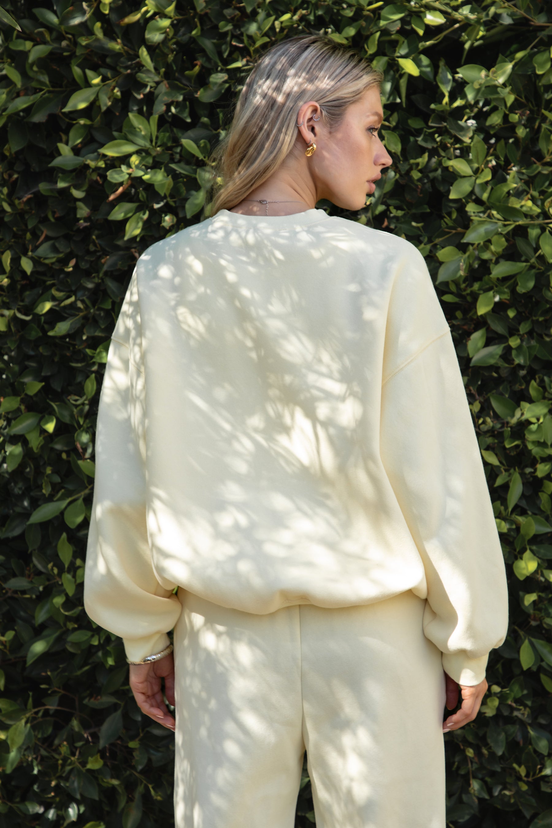 Woman wearing the TIC TOC TH28243 VARSITY pale yellow fleece crew neck pullover sweatshirt featuring dropped shoulders, ribbed trim, and plush fleece fabric with the matching bottoms standing against a green hedge.