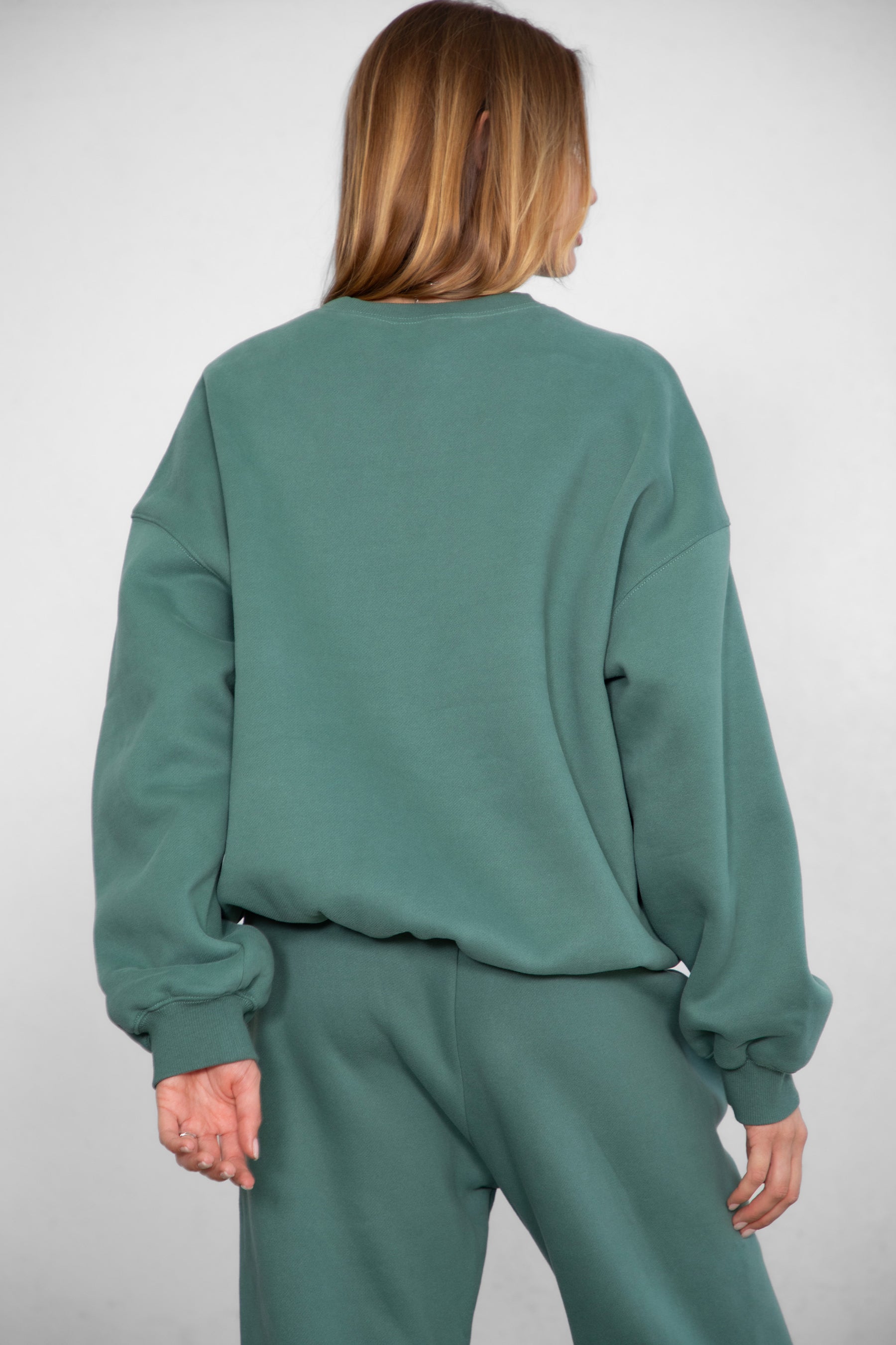 Woman wearing the TIC TOC TH28243 VARSITY pine green fleece crew neck pullover sweatshirt featuring dropped shoulders, ribbed trim, and plush fleece fabric with the matching bottoms