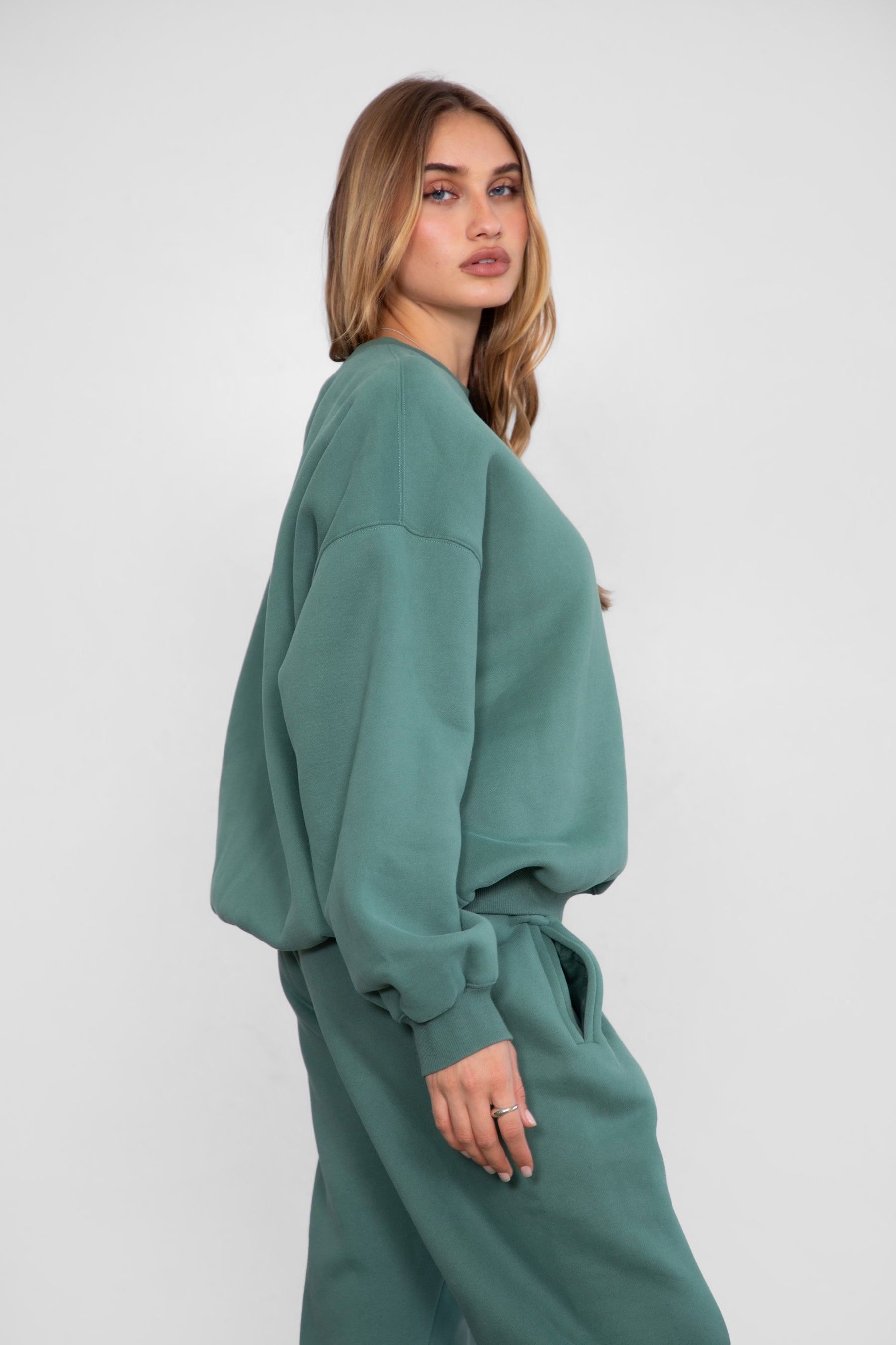 Woman wearing the TIC TOC TH28243 VARSITY pine green fleece crew neck pullover sweatshirt featuring dropped shoulders, ribbed trim, and plush fleece fabric with the matching bottoms