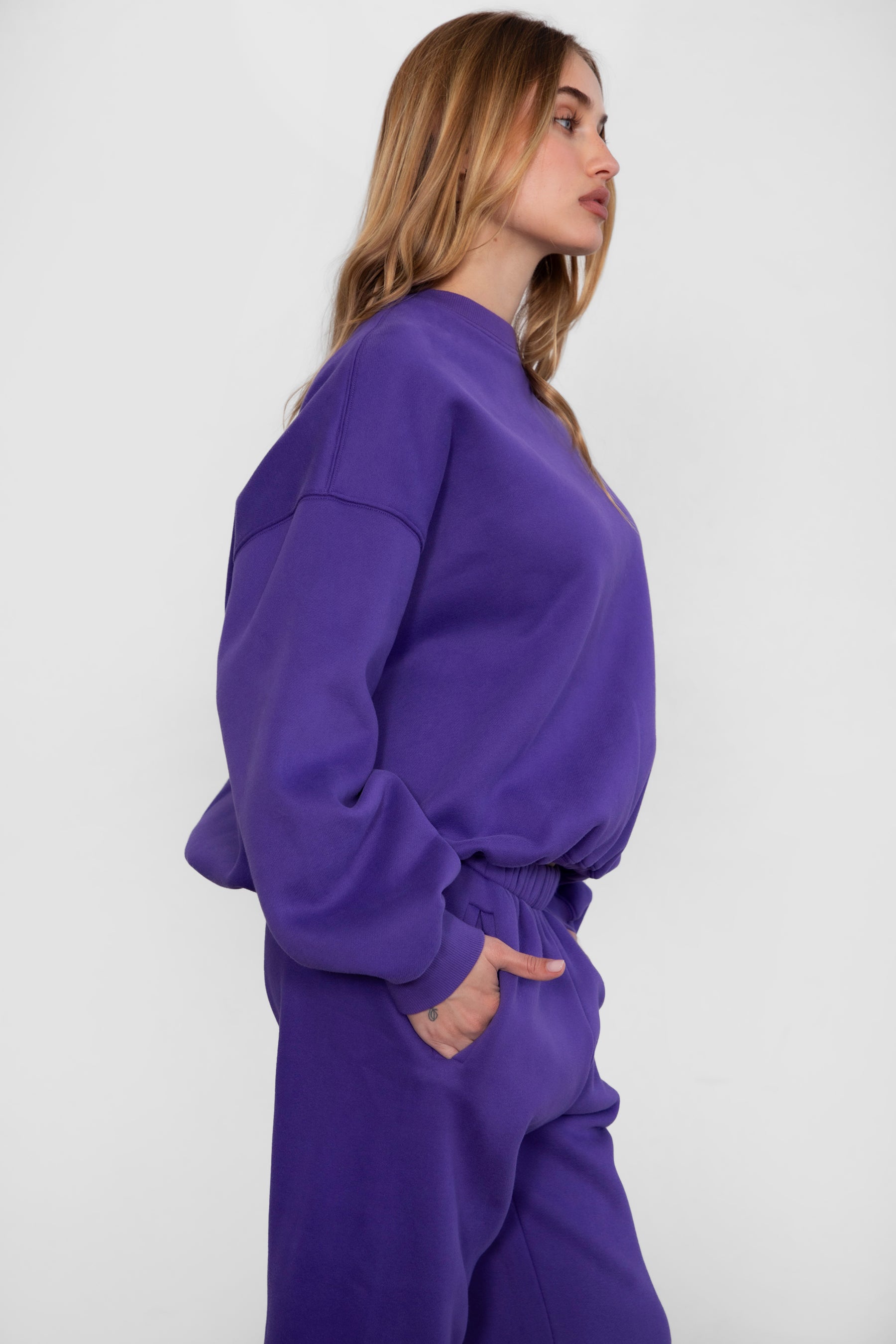 Woman wearing the TIC TOC TH28243 VARSITY purple fleece crew neck pullover sweatshirt featuring dropped shoulders, ribbed trim, and plush fleece fabric with the matching bottoms