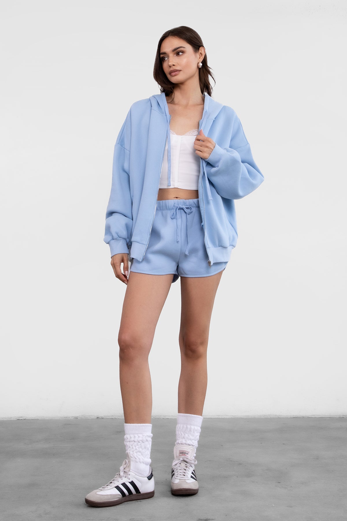 Woman wearing the TIC TOC TH29489 UNIVERSITY baby blue oversized loose fit zip up hoodie featuring dropped shoulders, ribbed hem, and plush fleece fabric with matching bottoms standing against a grey background.