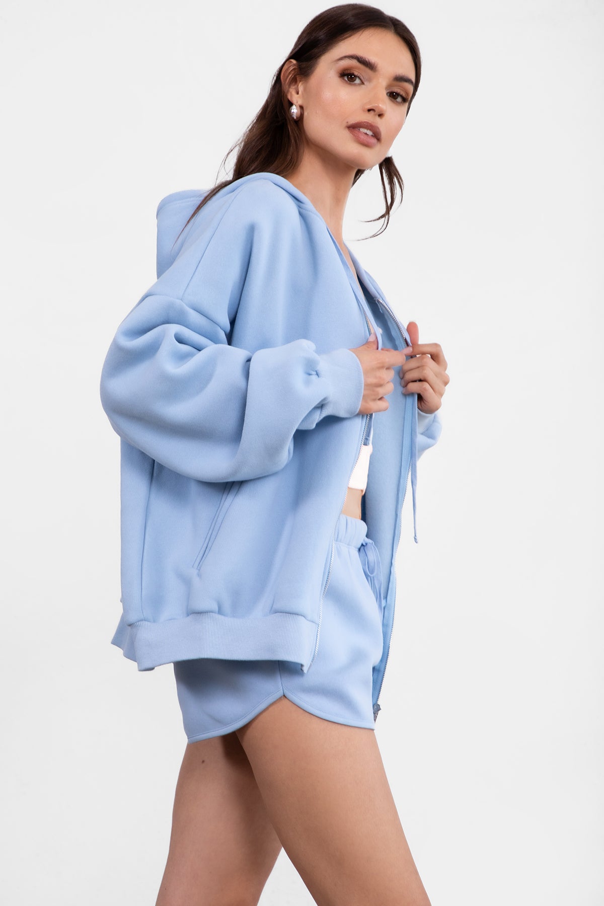 Woman wearing the TIC TOC TH29489 UNIVERSITY baby blue oversized loose fit zip up hoodie featuring dropped shoulders, ribbed hem, and plush fleece fabric with matching bottoms standing against a grey background.