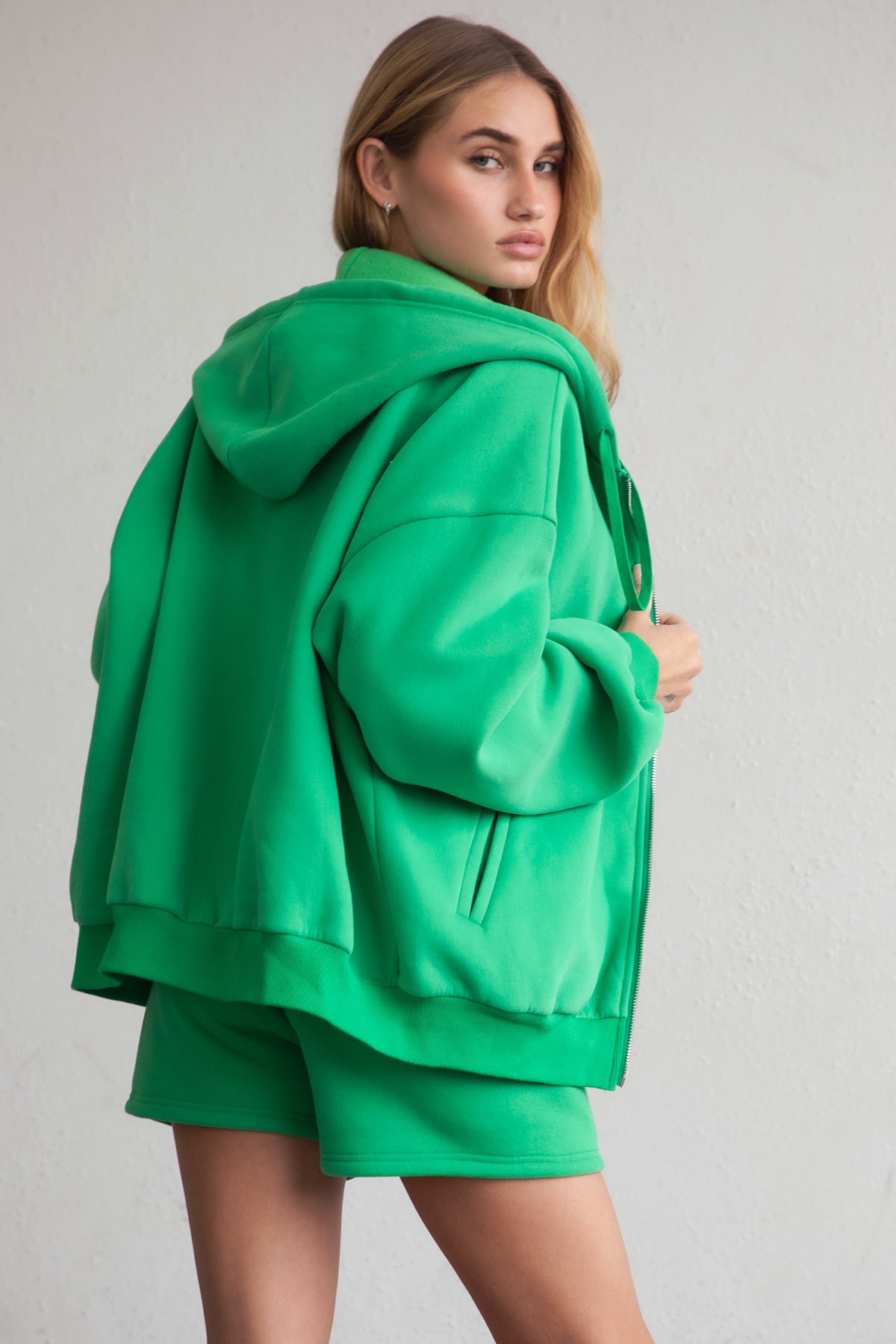 Woman wearing the TIC TOC TH29489 UNIVERSITY bright green oversized loose fit zip up hoodie featuring dropped shoulders, ribbed hem, and plush fleece fabric with matching bottoms standing against a grey background.