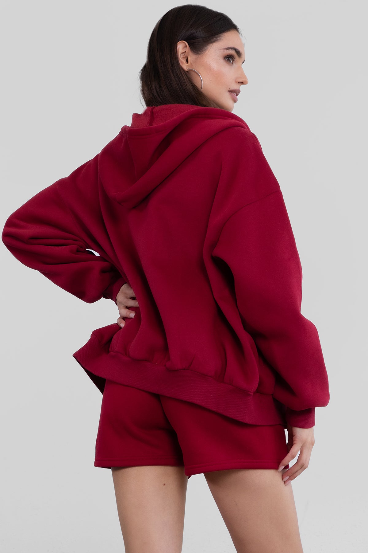 Woman wearing the TIC TOC TH29489 UNIVERSITY burgundy oversized loose fit zip up hoodie featuring dropped shoulders, ribbed hem, and plush fleece fabric with matching bottoms standing against a grey background.