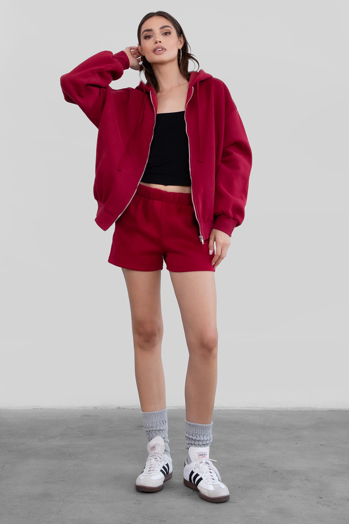 Woman wearing the TIC TOC TH29489 UNIVERSITY burgundy oversized loose fit zip up hoodie featuring dropped shoulders, ribbed hem, and plush fleece fabric with matching bottoms standing against a grey background.