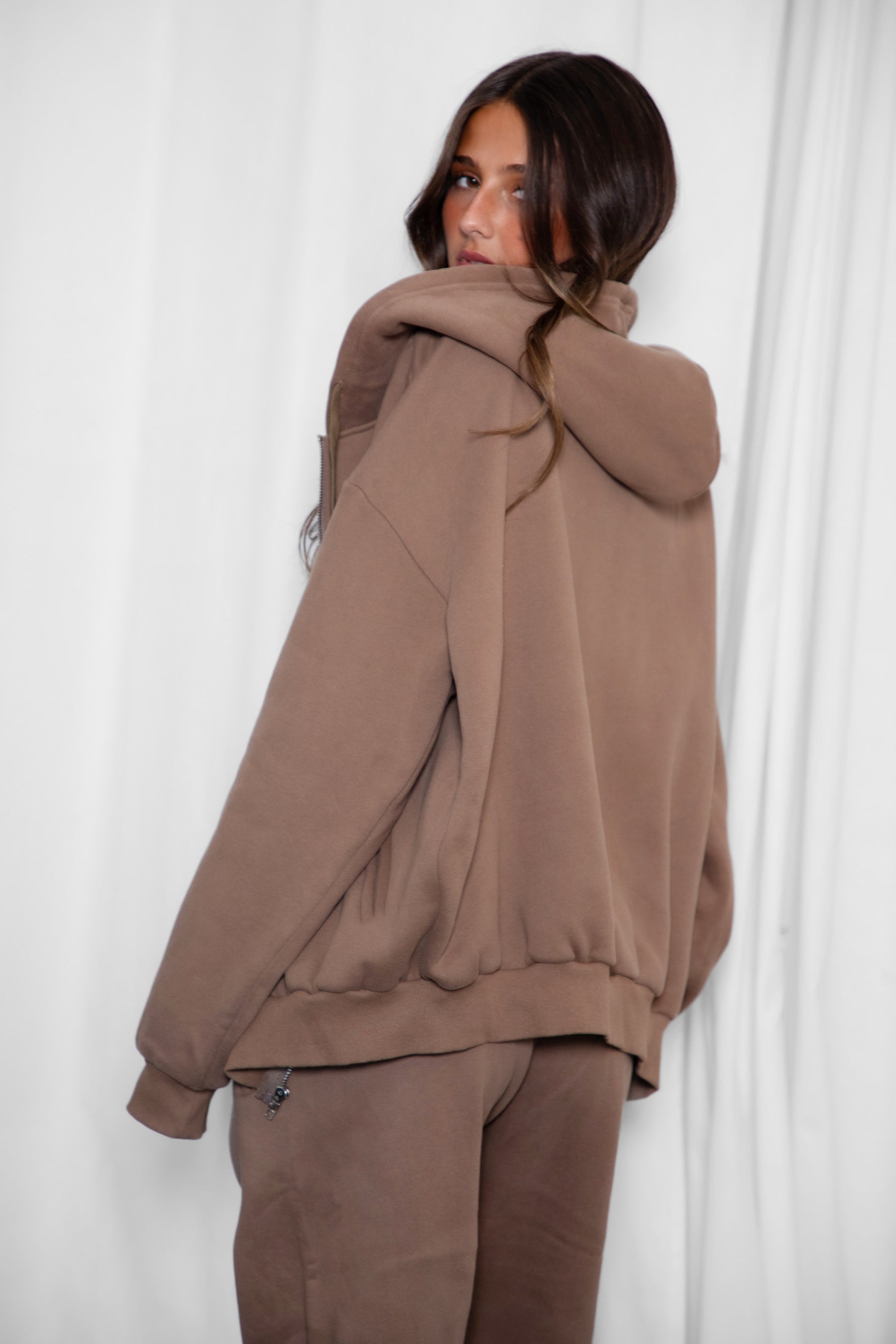 Woman wearing the TIC TOC TH29489 UNIVERSITY coffee oversized loose fit zip up hoodie featuring dropped shoulders, ribbed hem, and plush fleece fabric with matching bottoms standing against a grey background.