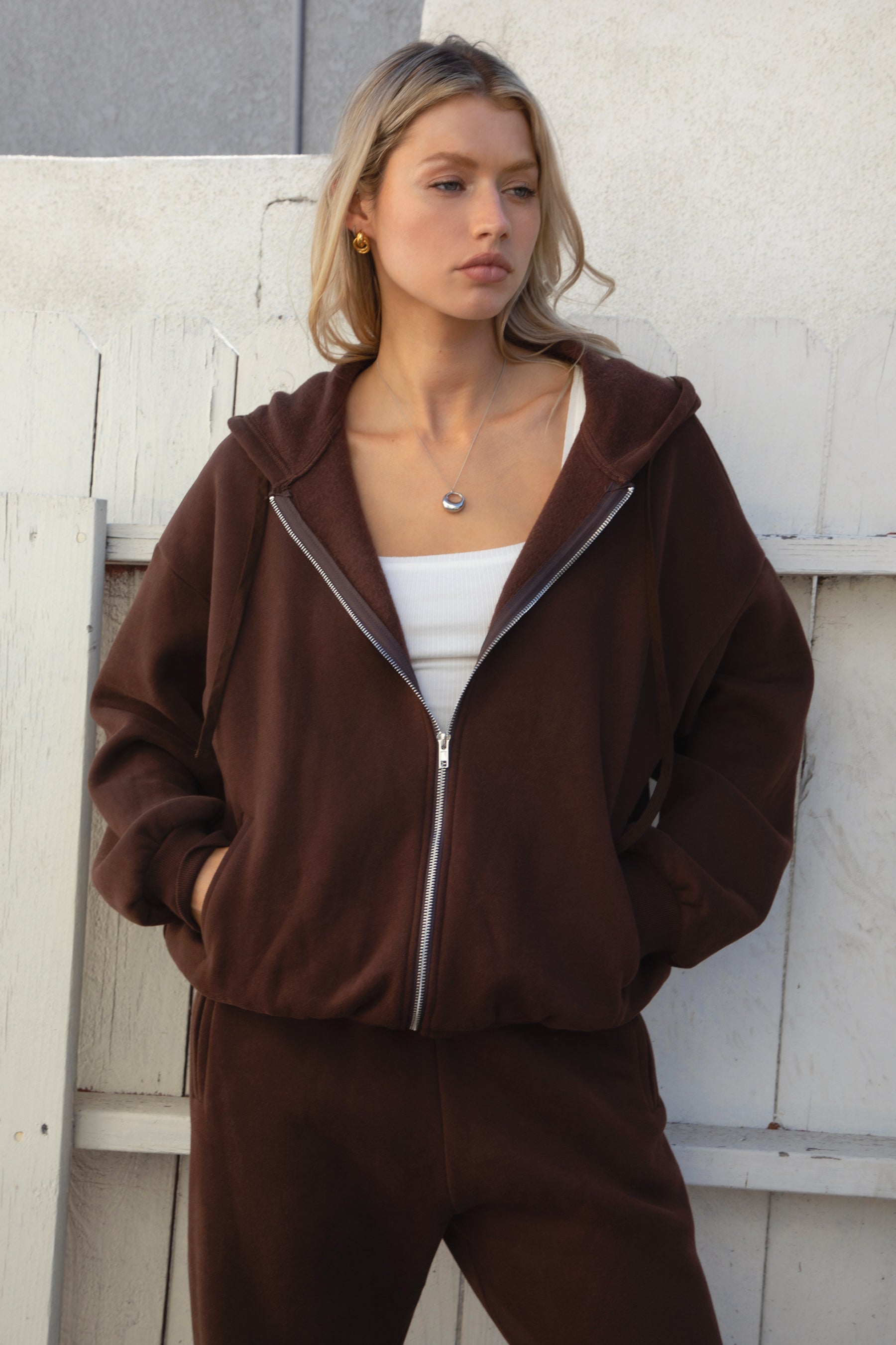 Woman wearing the TIC TOC TH29489 UNIVERSITY brown oversized loose fit zip up hoodie featuring dropped shoulders, ribbed hem, and plush fleece fabric with matching bottoms against a light-colored wall.