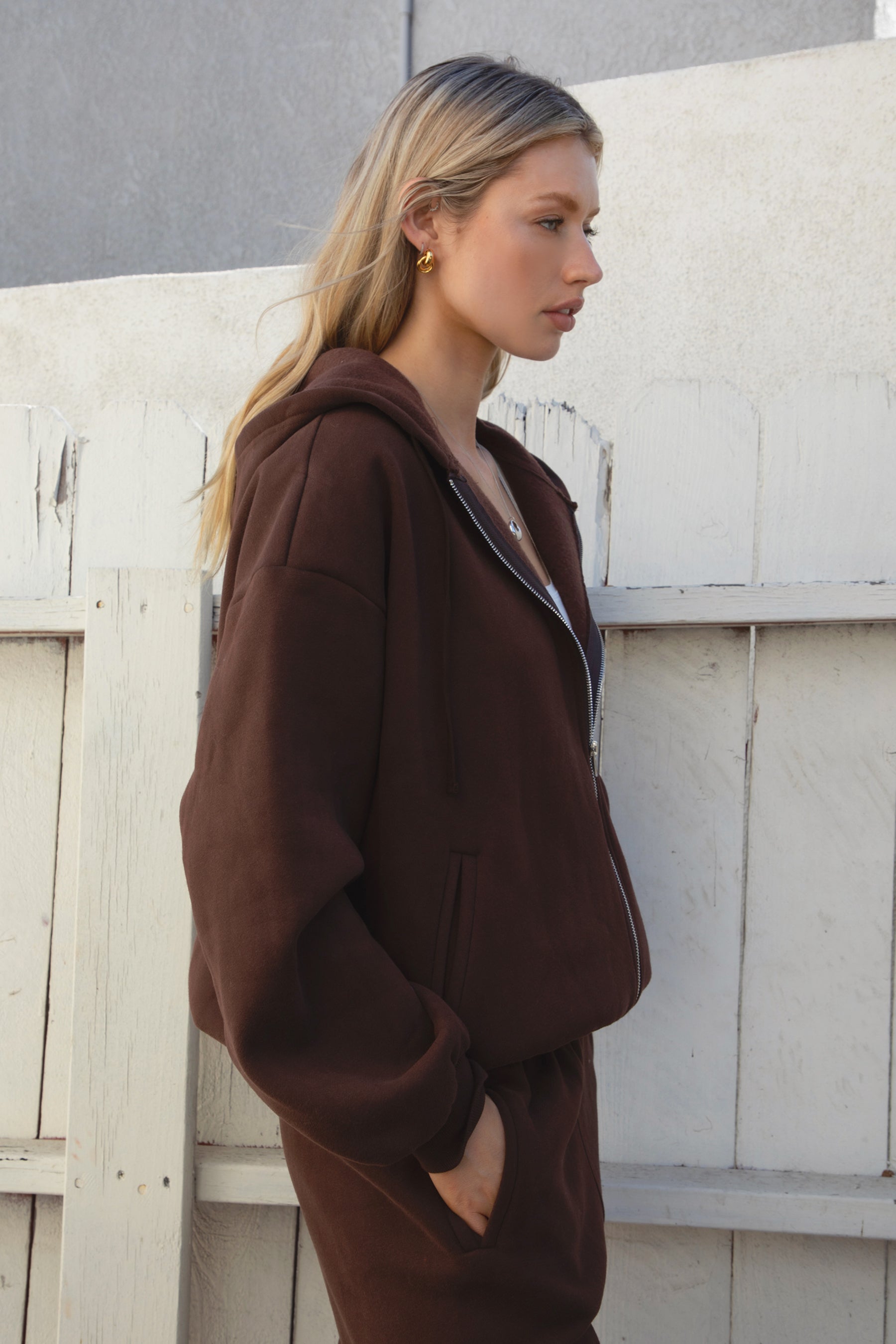 Woman wearing the TIC TOC TH29489 UNIVERSITY brown oversized loose fit zip up hoodie featuring dropped shoulders, ribbed hem, and plush fleece fabric with matching bottoms against a light-colored wall.