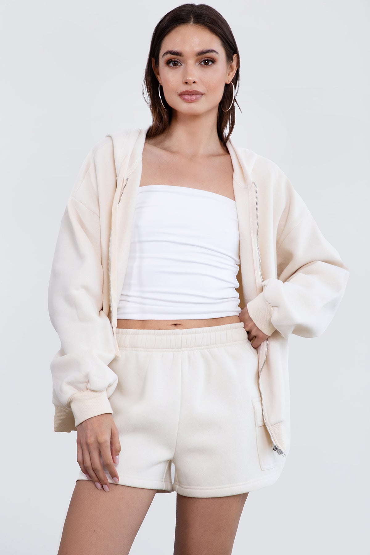 Woman wearing the TIC TOC TH29489 UNIVERSITY ecru oversized loose fit zip up hoodie featuring dropped shoulders, ribbed hem, and plush fleece fabric with matching bottoms standing against a grey background.