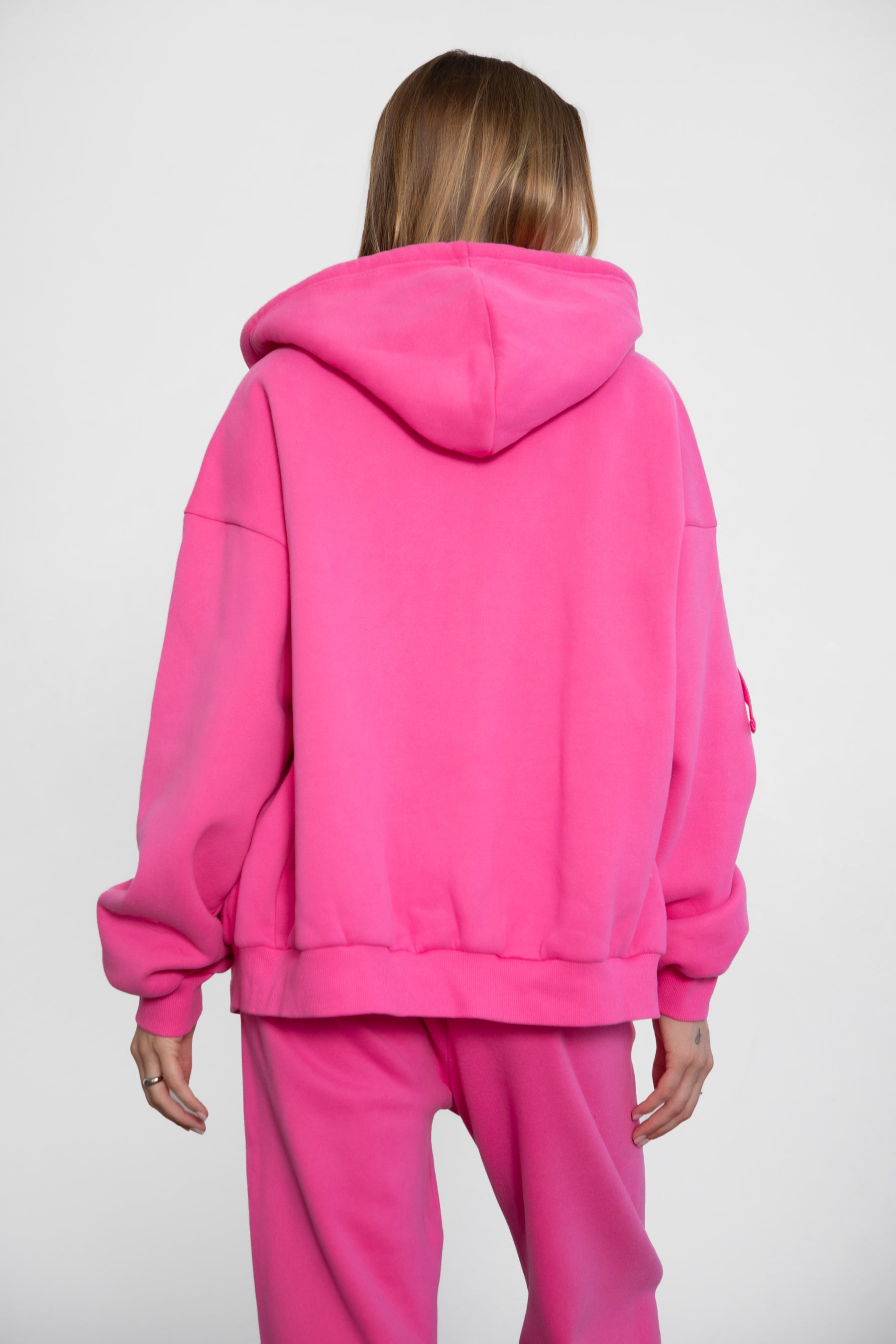 Woman wearing the TIC TOC TH29489 UNIVERSITY fuchsia oversized loose fit zip up hoodie featuring dropped shoulders, ribbed hem, and plush fleece fabric with matching bottoms standing against a grey background.