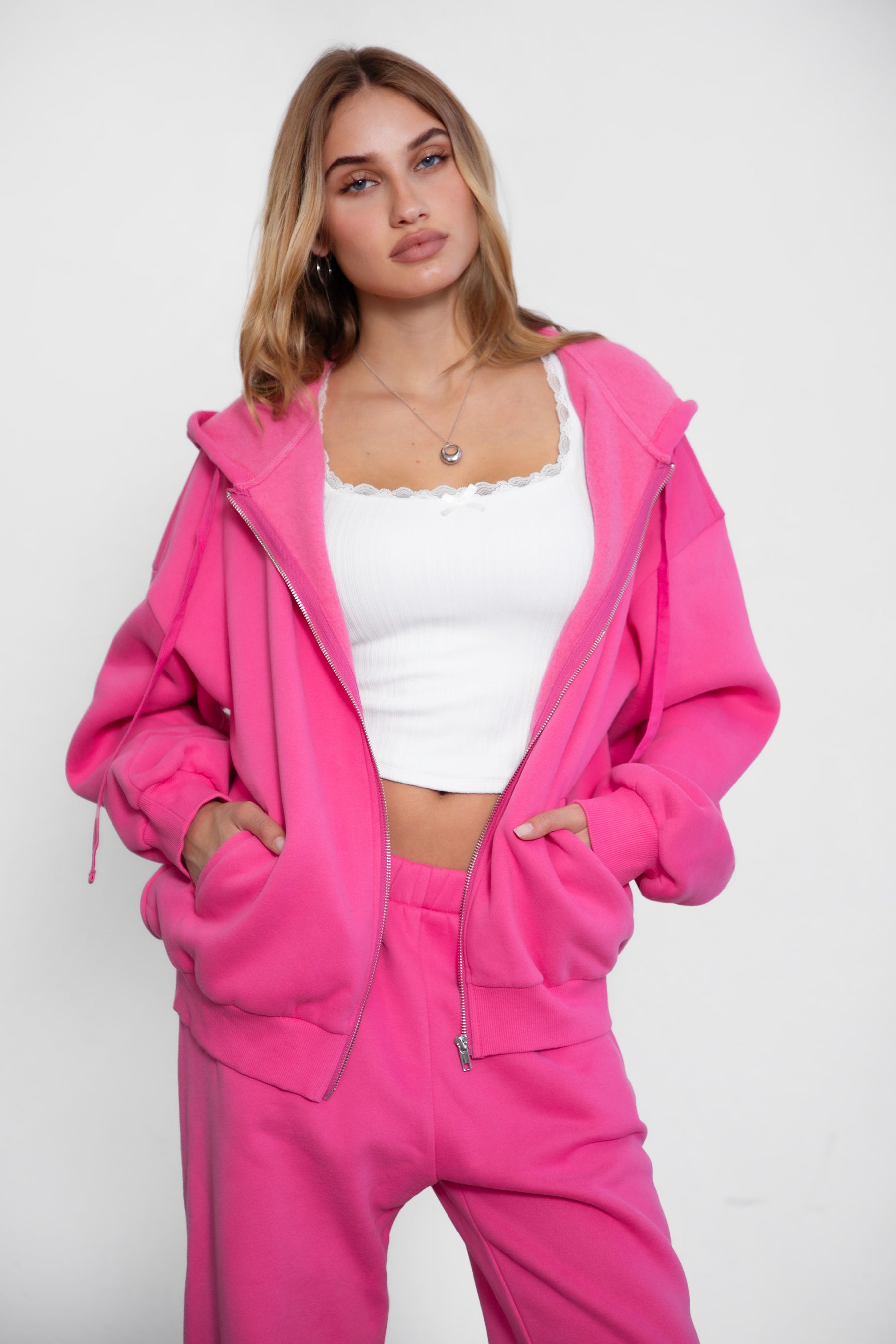 Woman wearing the TIC TOC TH29489 UNIVERSITY fuchsia oversized loose fit zip up hoodie featuring dropped shoulders, ribbed hem, and plush fleece fabric with matching bottoms standing against a grey background.
