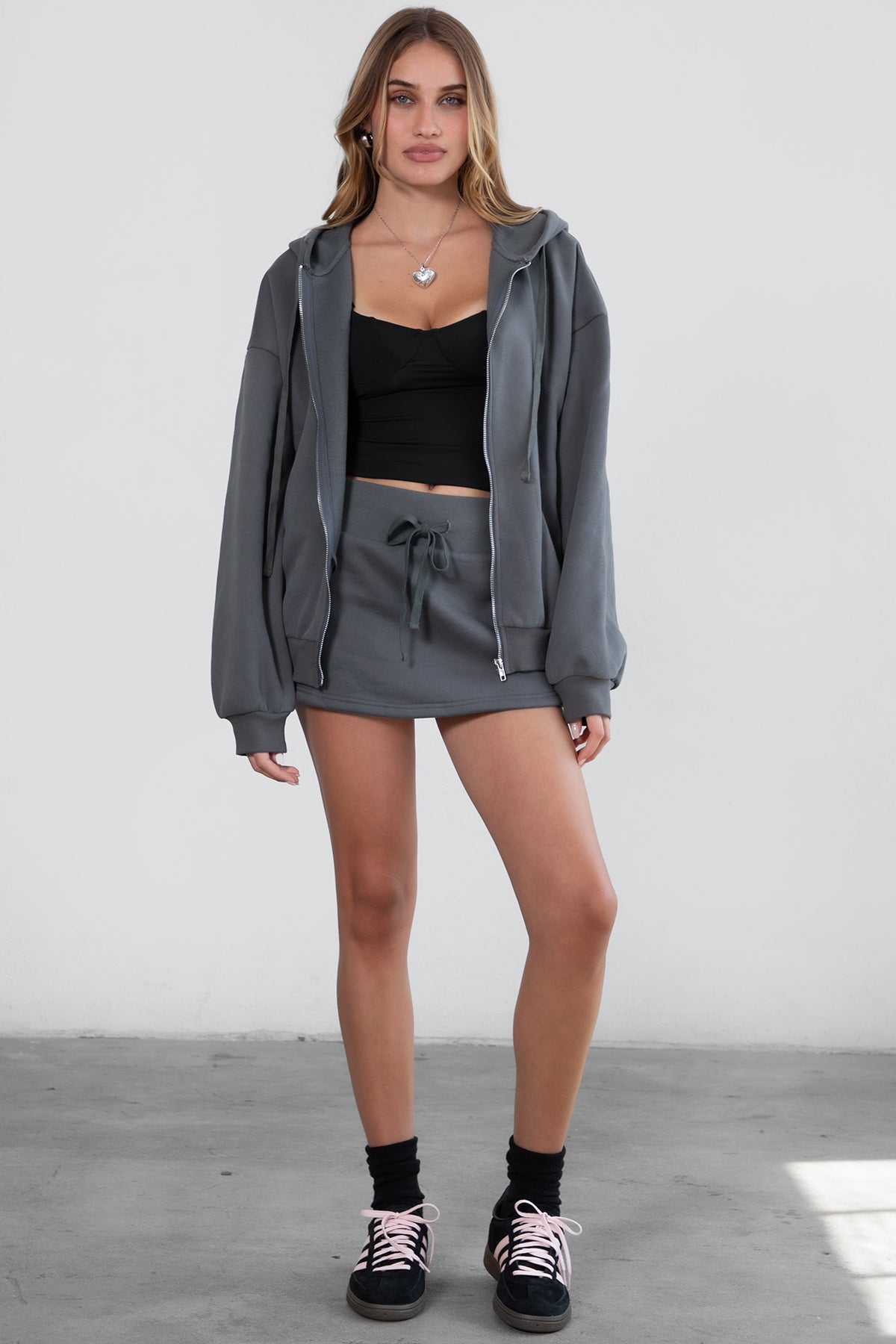 Woman wearing the TIC TOC TH29489 UNIVERSITY graphite oversized loose fit zip up hoodie featuring dropped shoulders, ribbed hem, and plush fleece fabric with matching bottoms standing against a grey background.