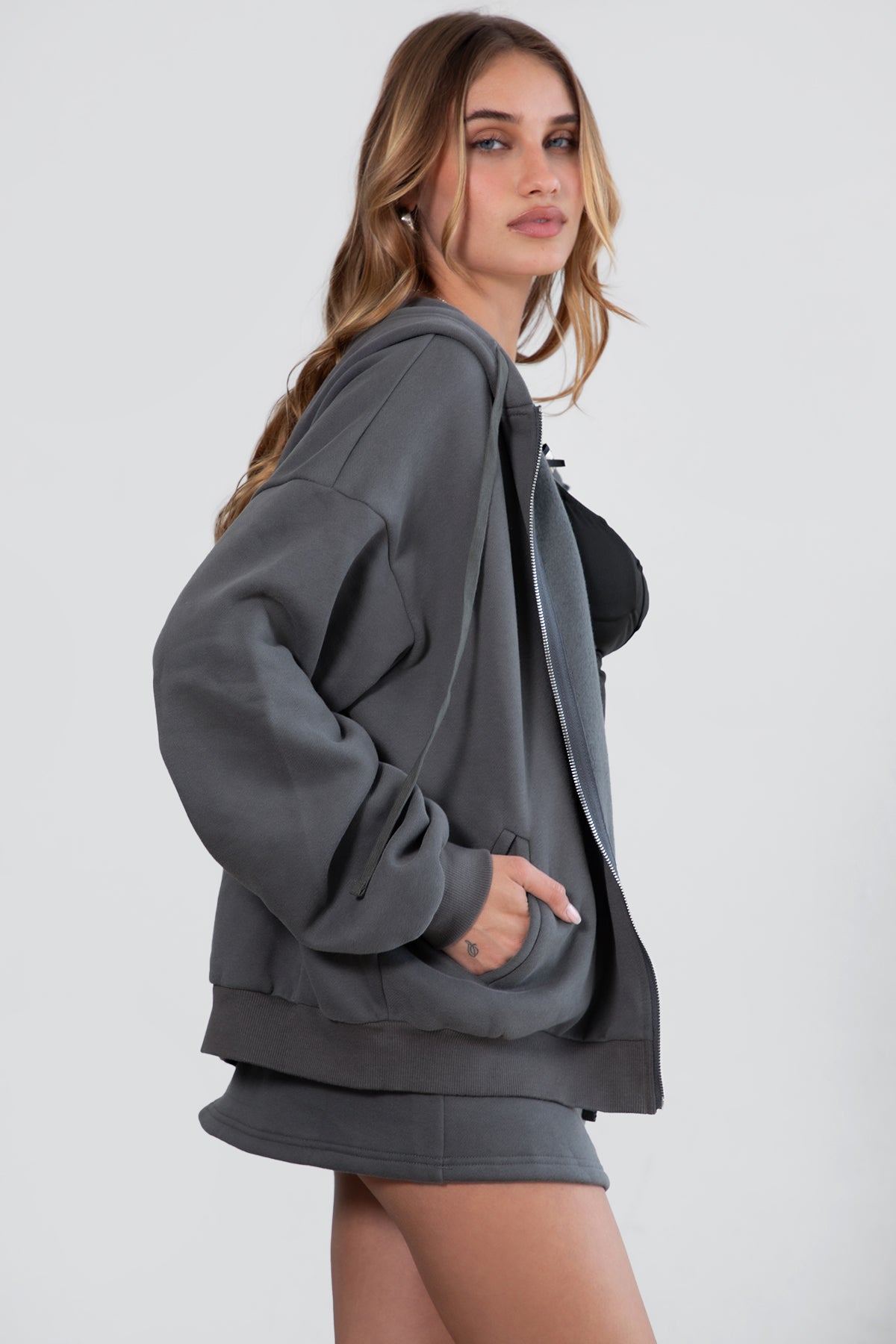 Woman wearing the TIC TOC TH29489 UNIVERSITY graphite oversized loose fit zip up hoodie featuring dropped shoulders, ribbed hem, and plush fleece fabric with matching bottoms standing against a grey background.