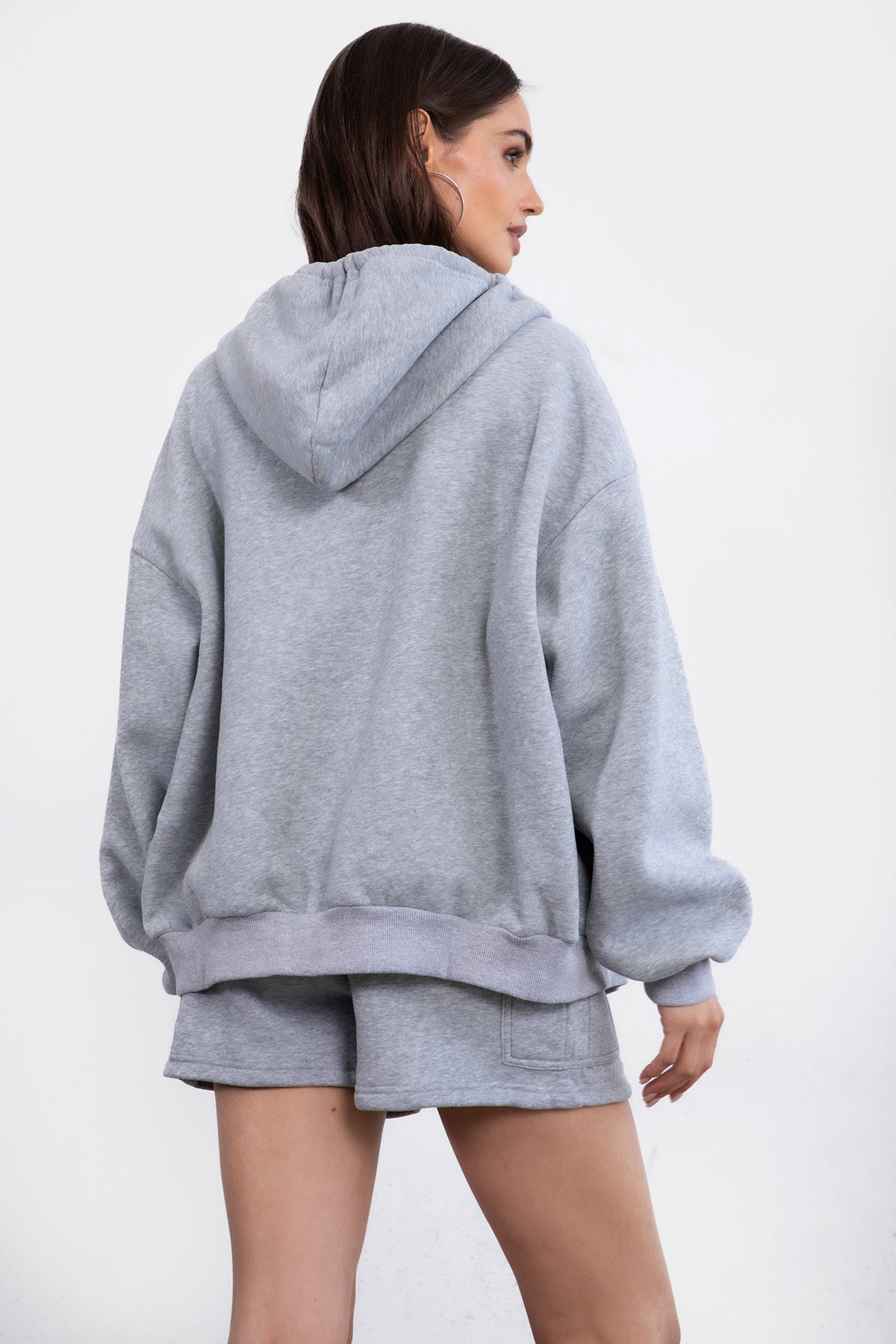 Woman wearing the TIC TOC TH29489 UNIVERSITY heather grey oversized loose fit zip up hoodie featuring dropped shoulders, ribbed hem, and plush fleece fabric with matching bottoms standing against a grey background.