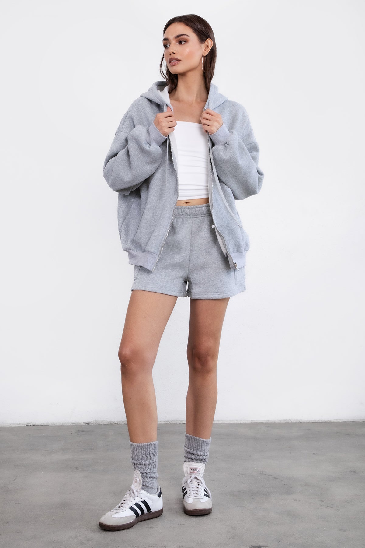 Woman wearing the TIC TOC TH29489 UNIVERSITY heather grey oversized loose fit zip up hoodie featuring dropped shoulders, ribbed hem, and plush fleece fabric with matching bottoms standing against a grey background.