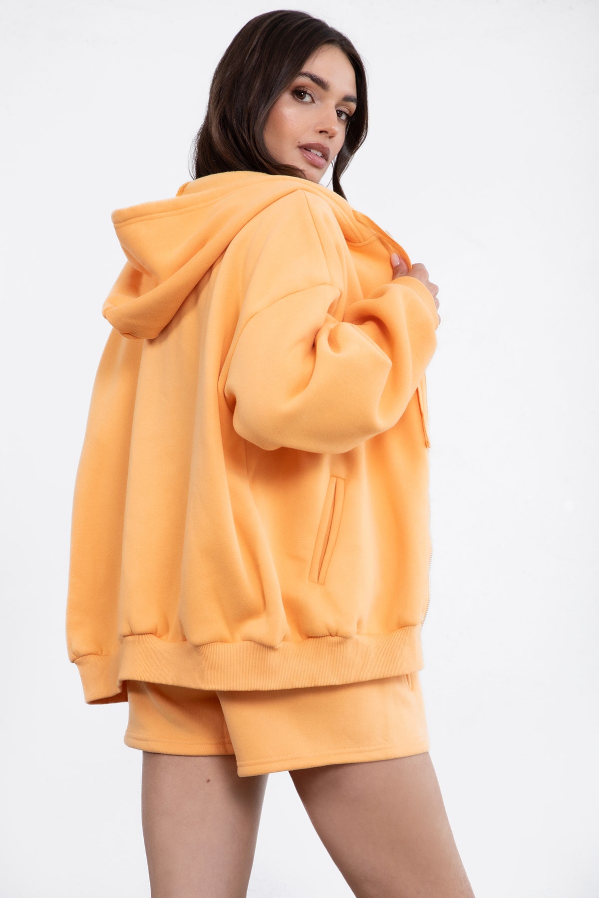 Woman wearing the TIC TOC TH29489 UNIVERSITY orange oversized loose fit zip up hoodie featuring dropped shoulders, ribbed hem, and plush fleece fabric with matching bottoms standing against a grey background.