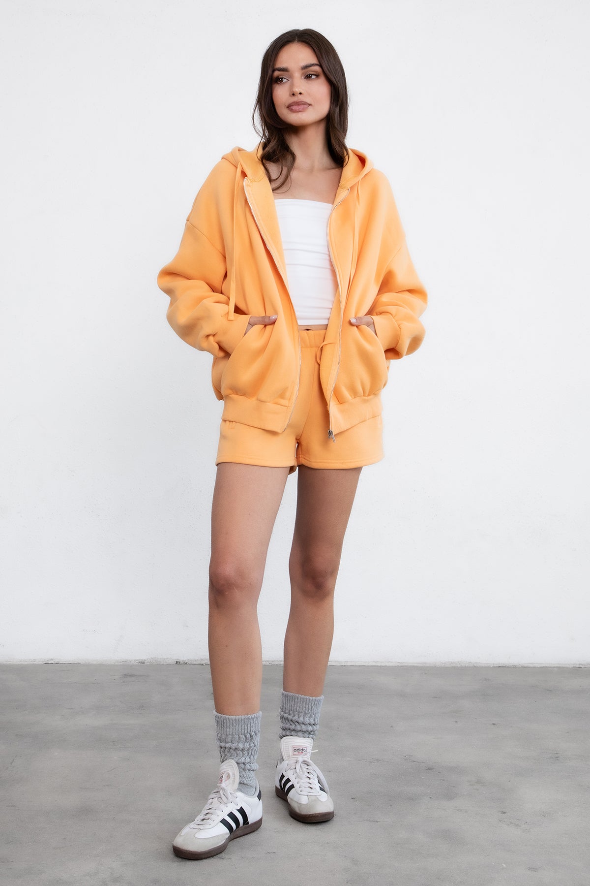 Woman wearing the TIC TOC TH29489 UNIVERSITY orange oversized loose fit zip up hoodie featuring dropped shoulders, ribbed hem, and plush fleece fabric with matching bottoms standing against a grey background.