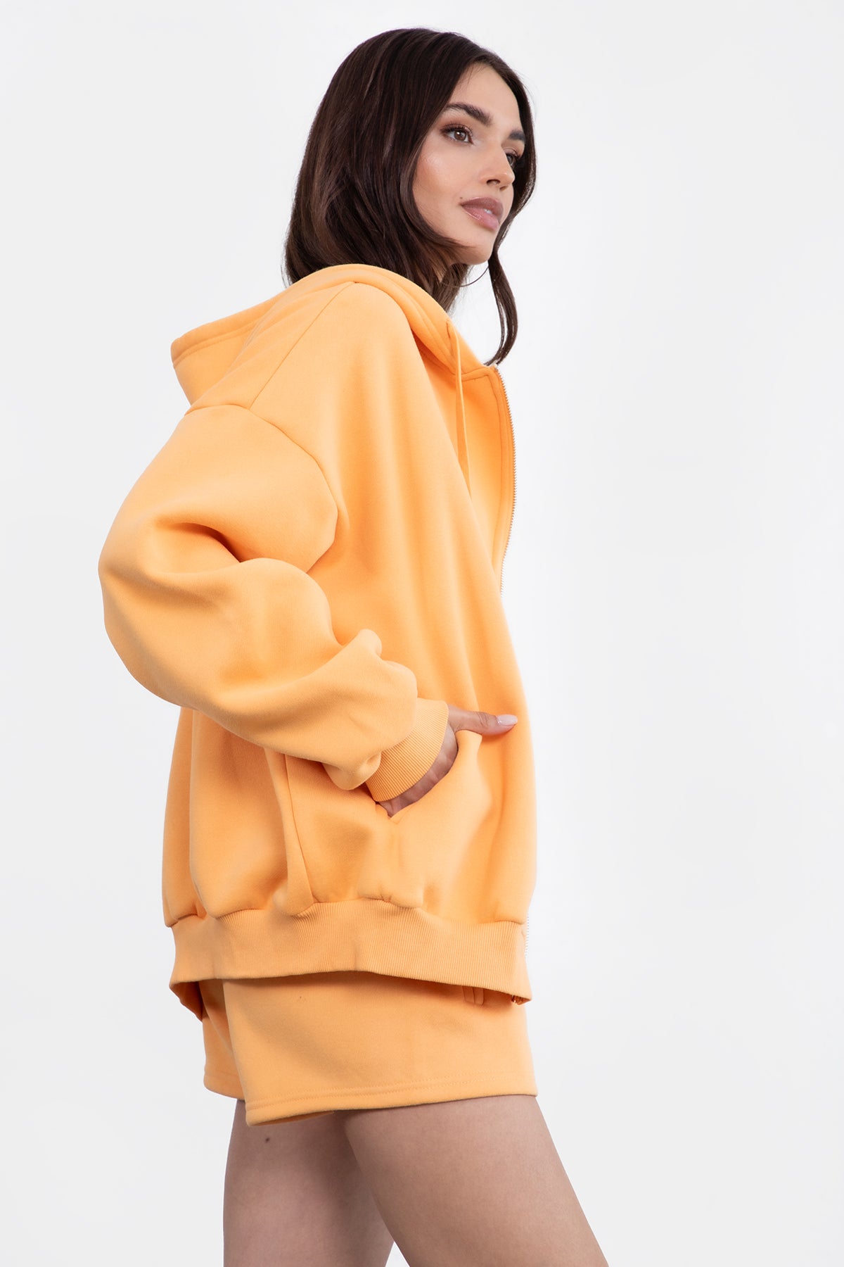 Woman wearing the TIC TOC TH29489 UNIVERSITY orange oversized loose fit zip up hoodie featuring dropped shoulders, ribbed hem, and plush fleece fabric with matching bottoms standing against a grey background.