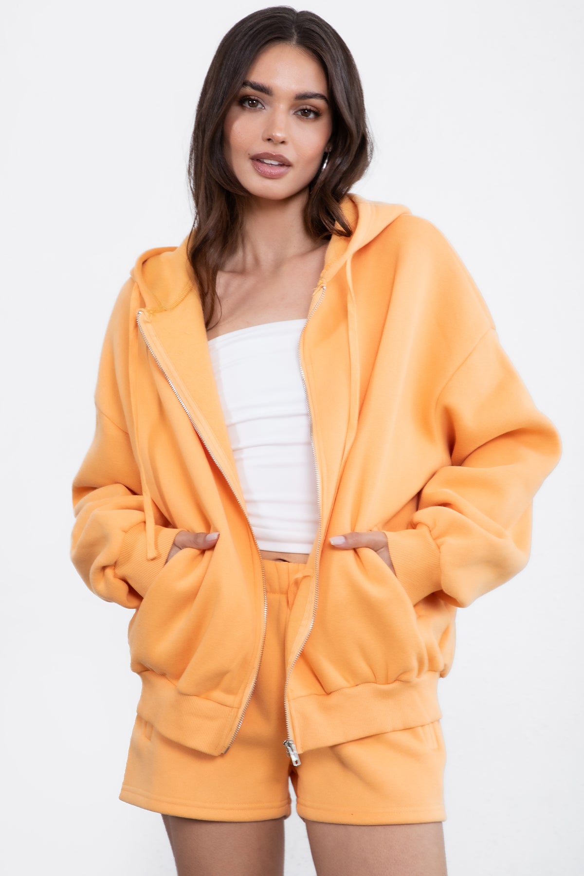 Woman wearing the TIC TOC TH29489 UNIVERSITY orange oversized loose fit zip up hoodie featuring dropped shoulders, ribbed hem, and plush fleece fabric with matching bottoms standing against a grey background.
