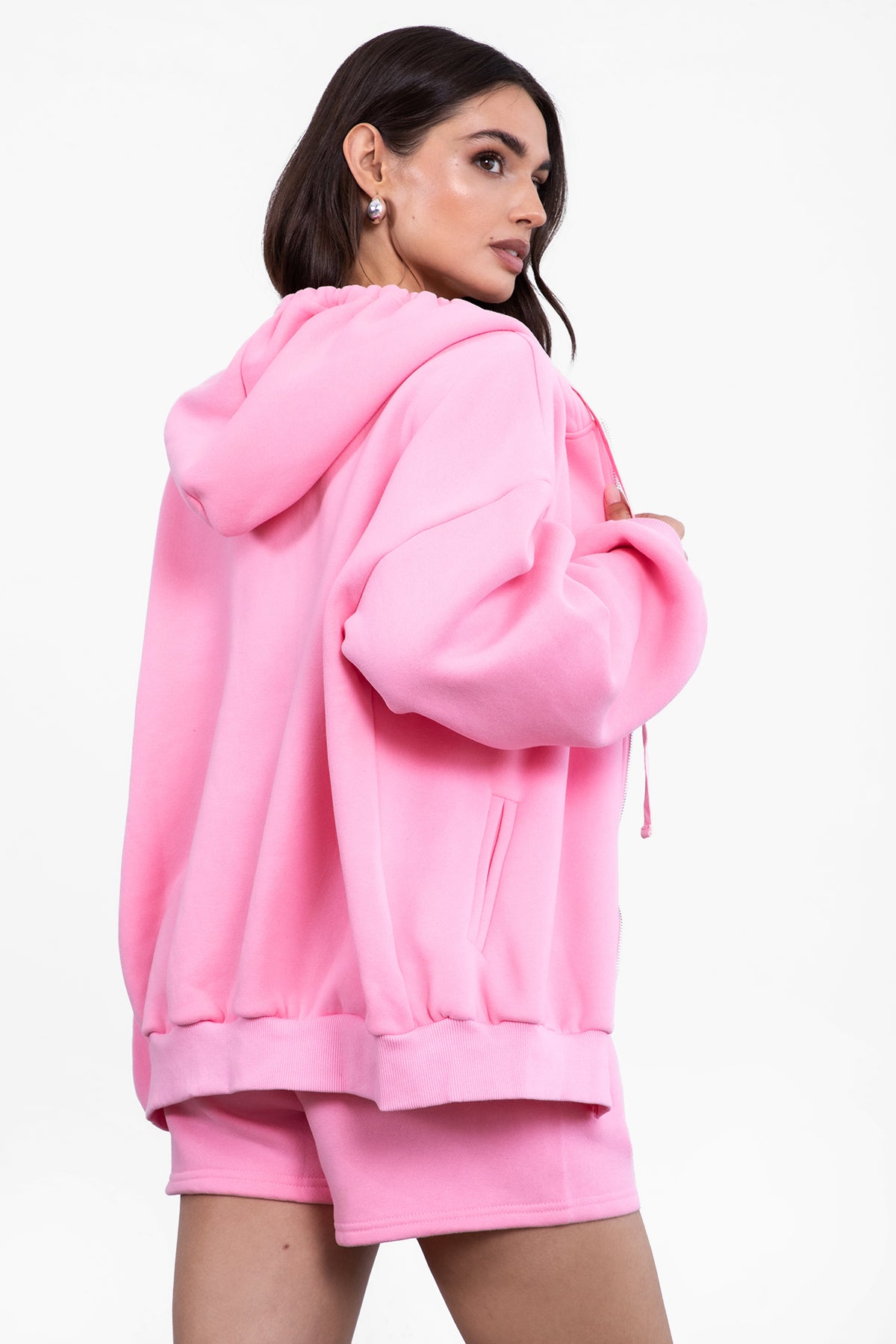 Woman wearing the TIC TOC TH29489 UNIVERSITY pink oversized loose fit zip up hoodie featuring dropped shoulders, ribbed hem, and plush fleece fabric with matching bottoms standing against a grey background.