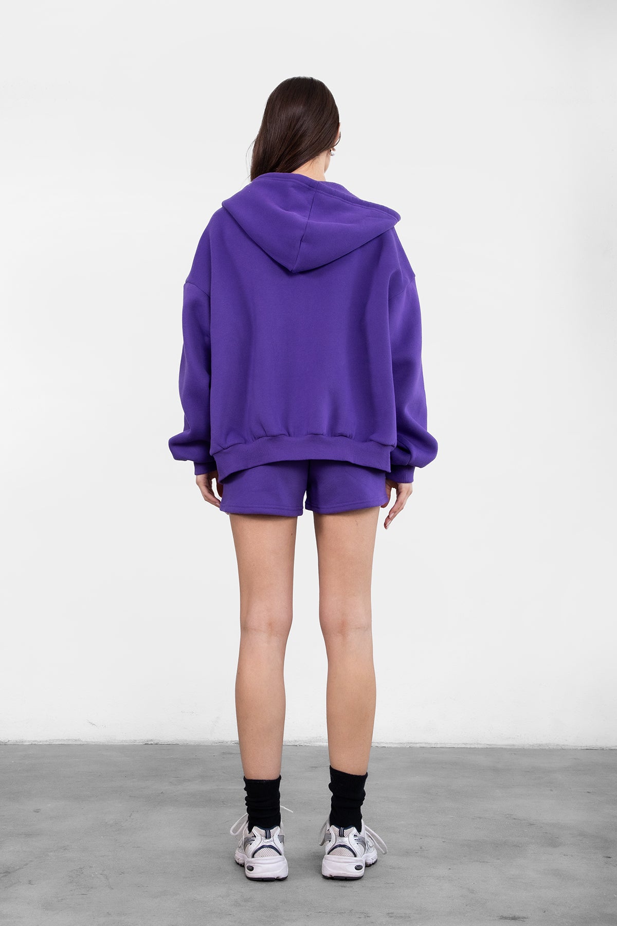 Woman wearing the TIC TOC TH29489 UNIVERSITY purple oversized loose fit zip up hoodie featuring dropped shoulders, ribbed hem, and plush fleece fabric with matching bottoms standing against a grey background.