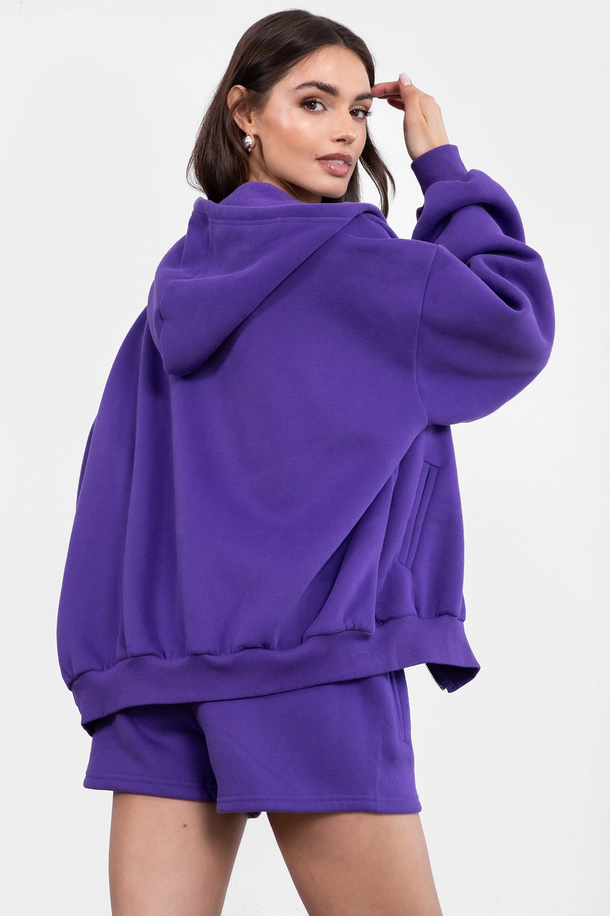 Woman wearing the TIC TOC TH29489 UNIVERSITY purple oversized loose fit zip up hoodie featuring dropped shoulders, ribbed hem, and plush fleece fabric with matching bottoms standing against a grey background.