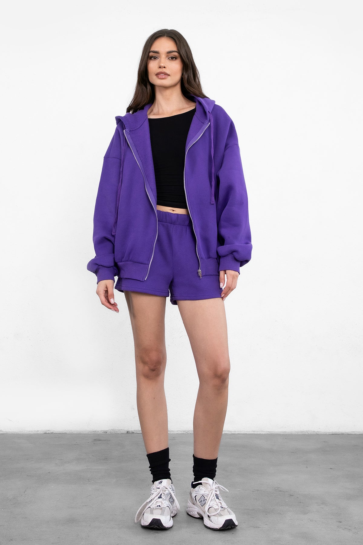 Woman wearing the TIC TOC TH29489 UNIVERSITY purple oversized loose fit zip up hoodie featuring dropped shoulders, ribbed hem, and plush fleece fabric with matching bottoms standing against a grey background.