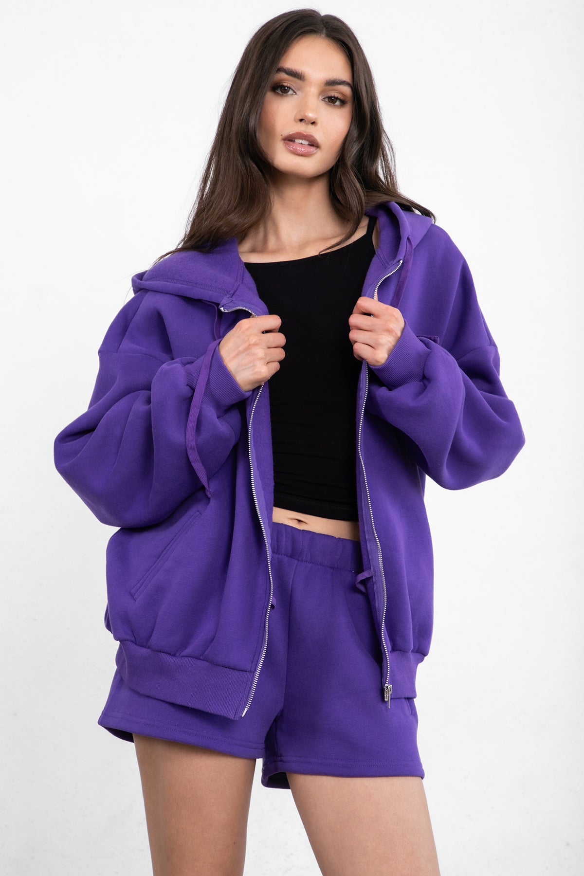 Woman wearing the TIC TOC TH29489 UNIVERSITY purple oversized loose fit zip up hoodie featuring dropped shoulders, ribbed hem, and plush fleece fabric with matching bottoms standing against a grey background.