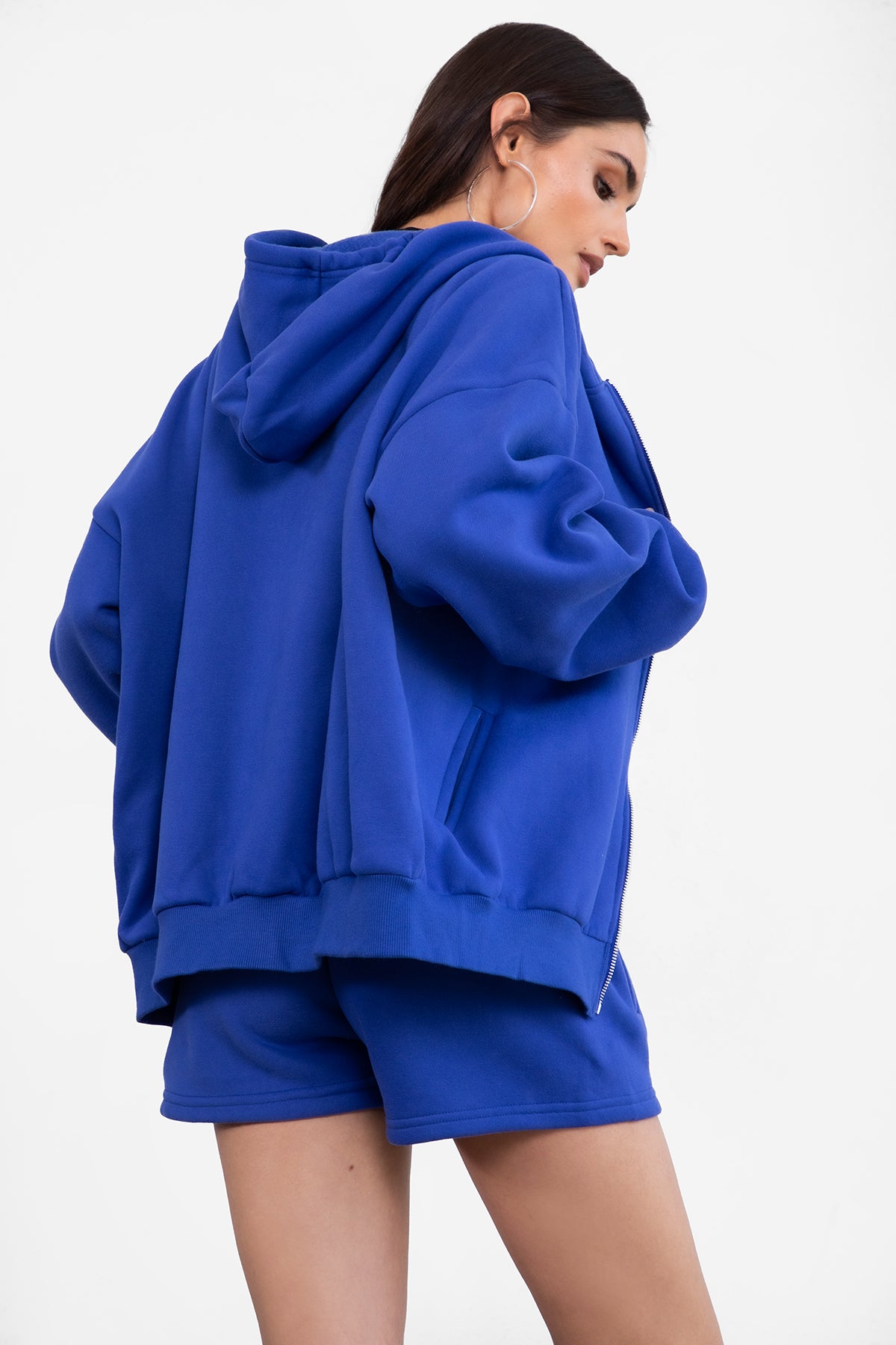 Woman wearing the TIC TOC TH29489 UNIVERSITY royal blue oversized loose fit zip up hoodie featuring dropped shoulders, ribbed hem, and plush fleece fabric with matching bottoms standing against a grey background.