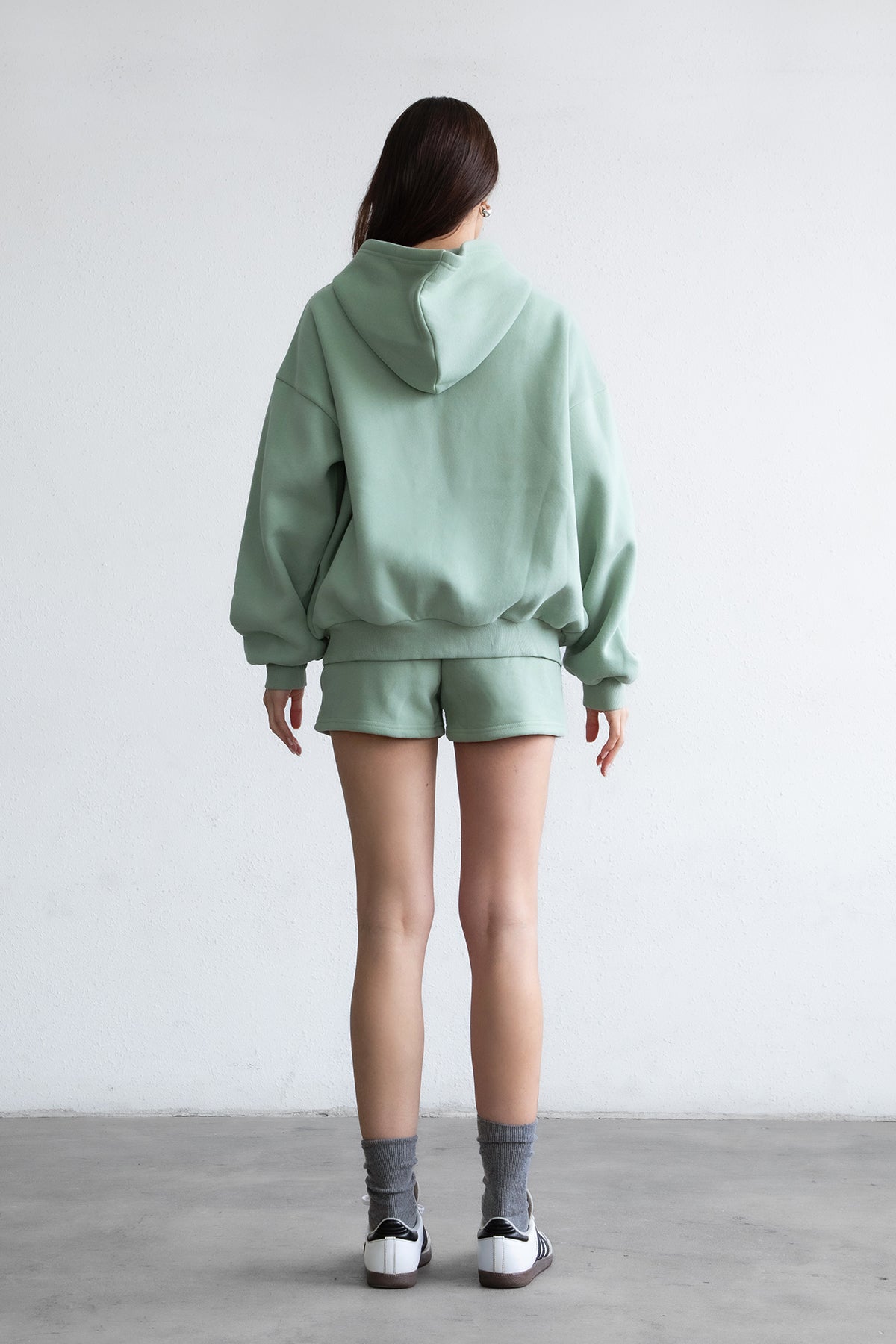Woman wearing the TIC TOC TH29489 UNIVERSITY sage oversized loose fit zip up hoodie featuring dropped shoulders, ribbed hem, and plush fleece fabric with matching bottoms standing against a grey background.