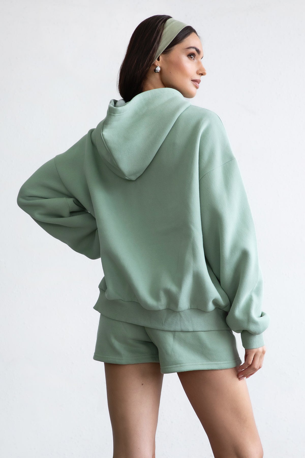 Woman wearing the TIC TOC TH29489 UNIVERSITY sage oversized loose fit zip up hoodie featuring dropped shoulders, ribbed hem, and plush fleece fabric with matching bottoms standing against a grey background.