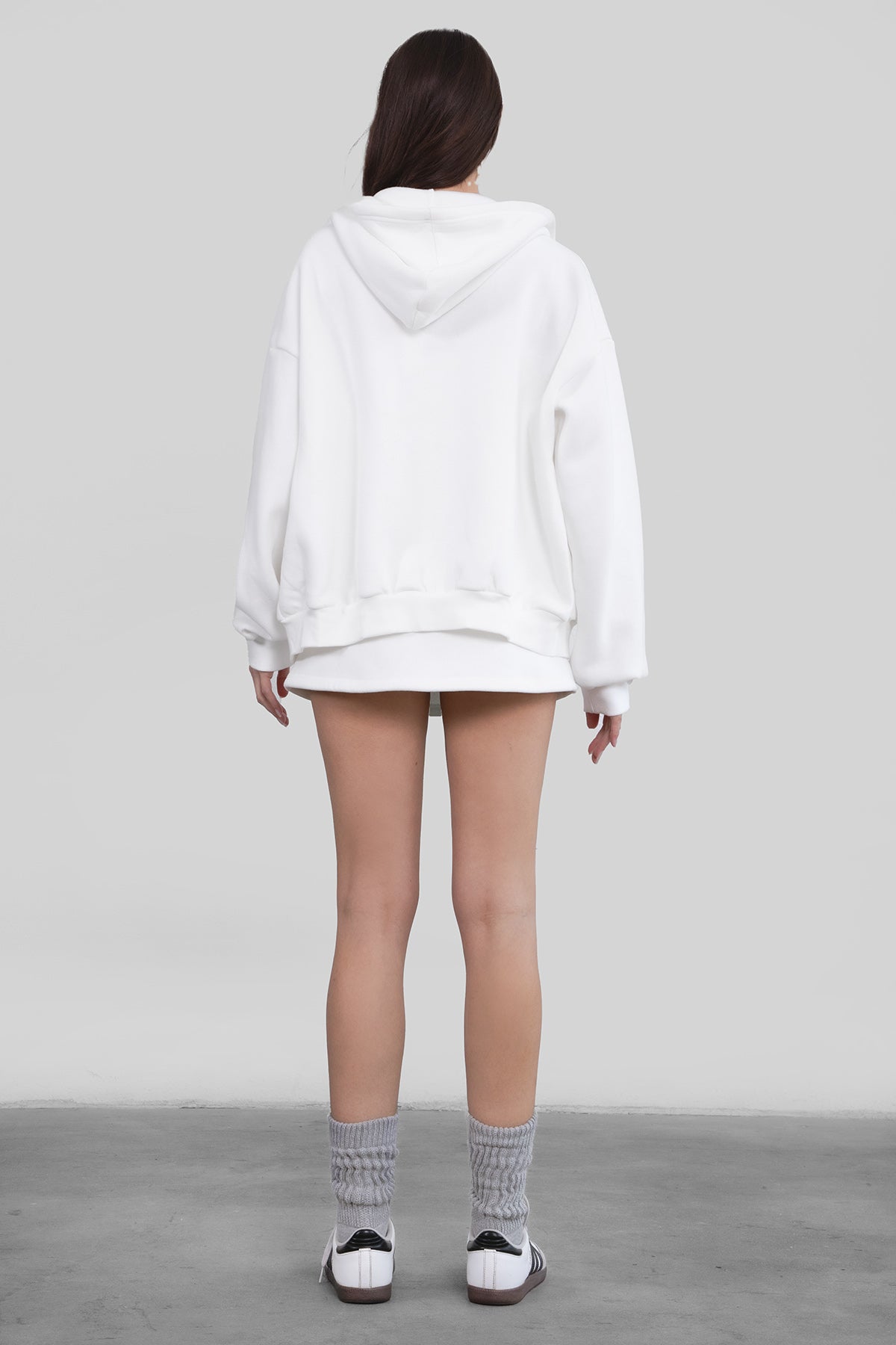 Woman wearing the TIC TOC TH29489 UNIVERSITY white oversized loose fit zip up hoodie featuring dropped shoulders, ribbed hem, and plush fleece fabric with matching bottoms standing against a grey background.