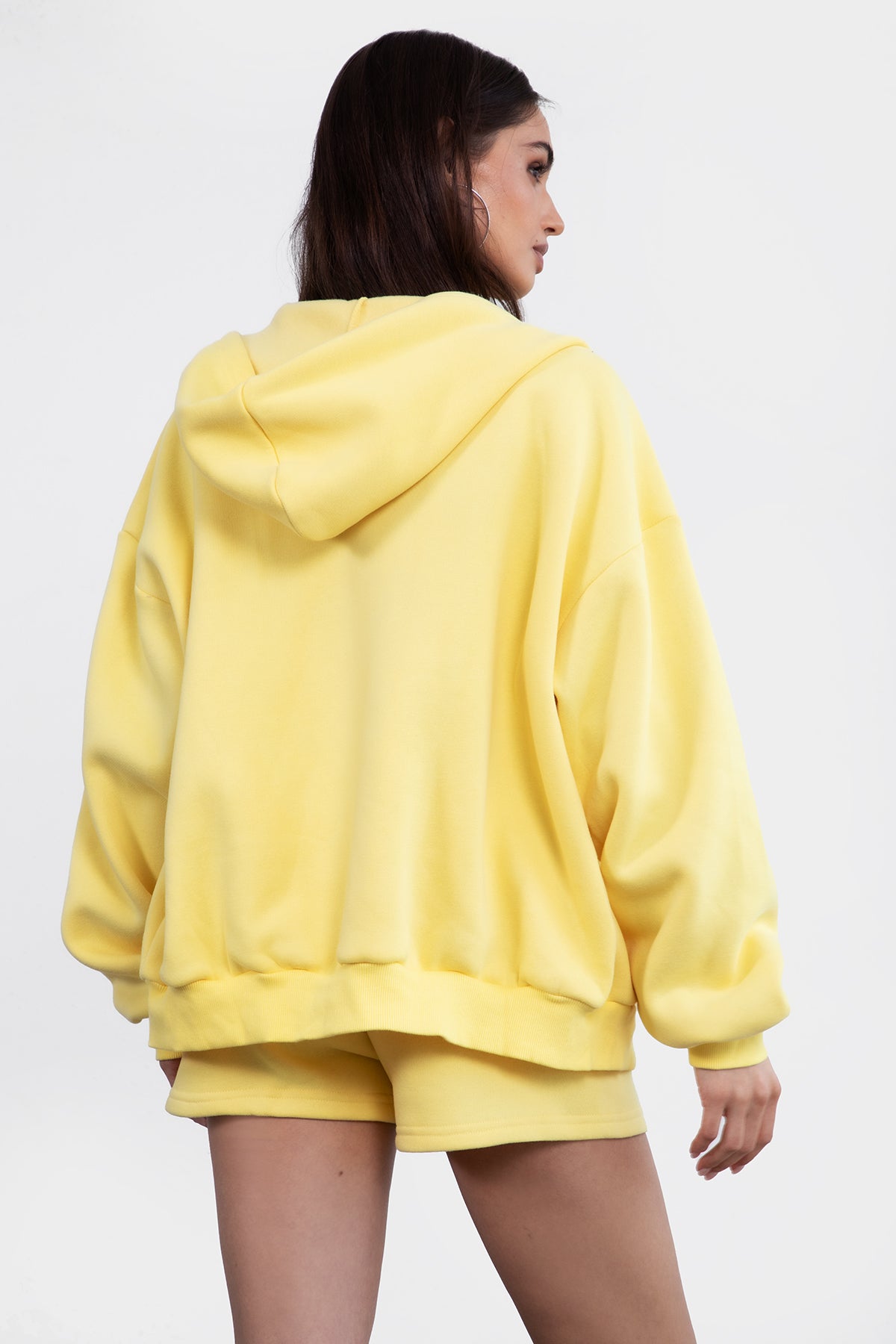 Woman wearing the TIC TOC TH29489 UNIVERSITY yellow oversized loose fit zip up hoodie featuring dropped shoulders, ribbed hem, and plush fleece fabric with matching bottoms standing against a grey background.
