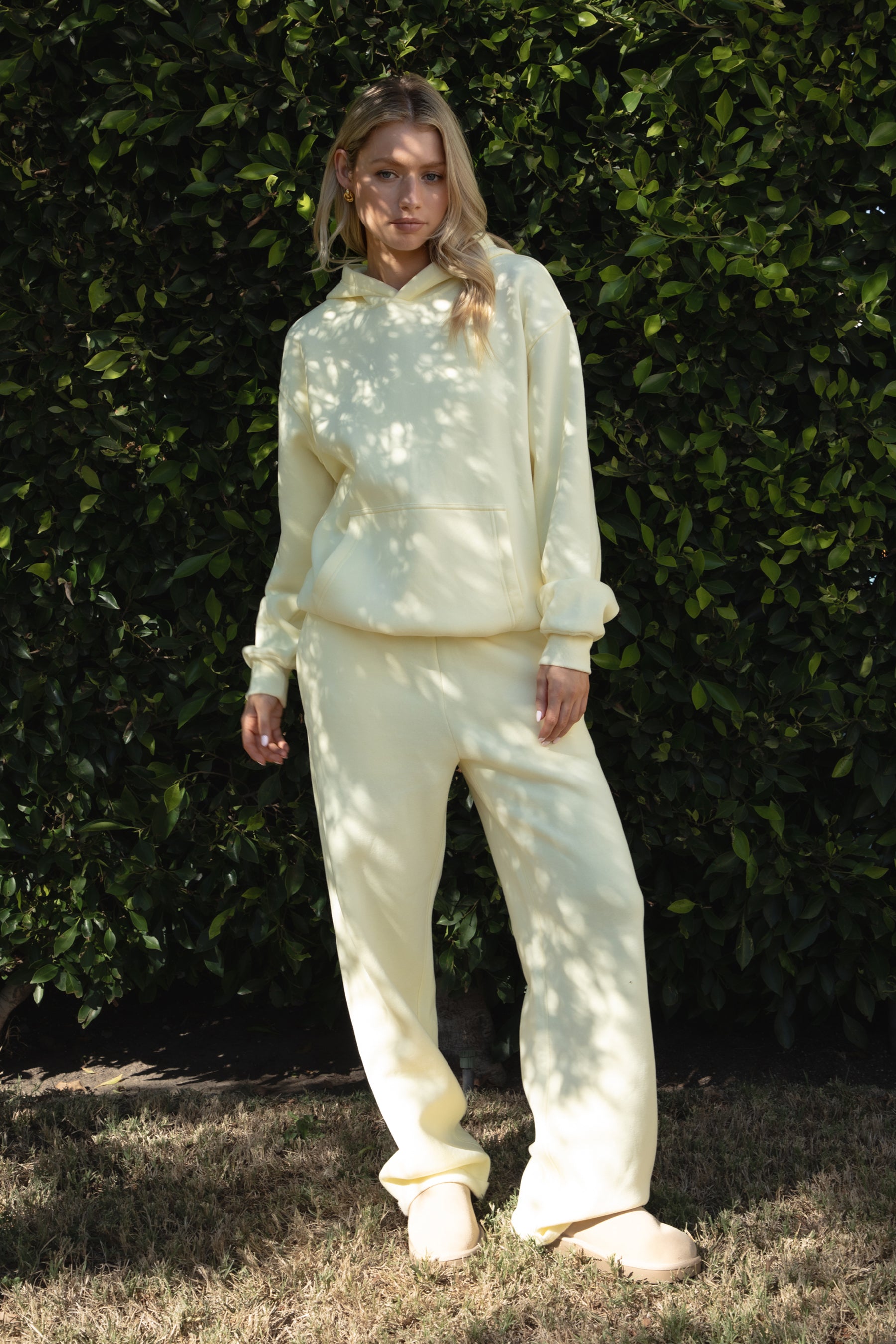Person wearing a yellow colored hoodie and pants standing against a green hedge.