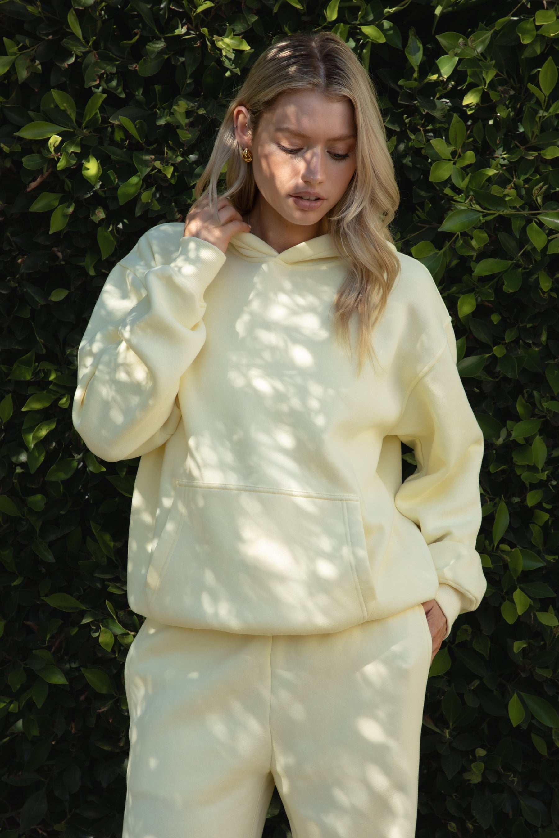 Person wearing a yellow colored hoodie and pants standing against a green hedge.