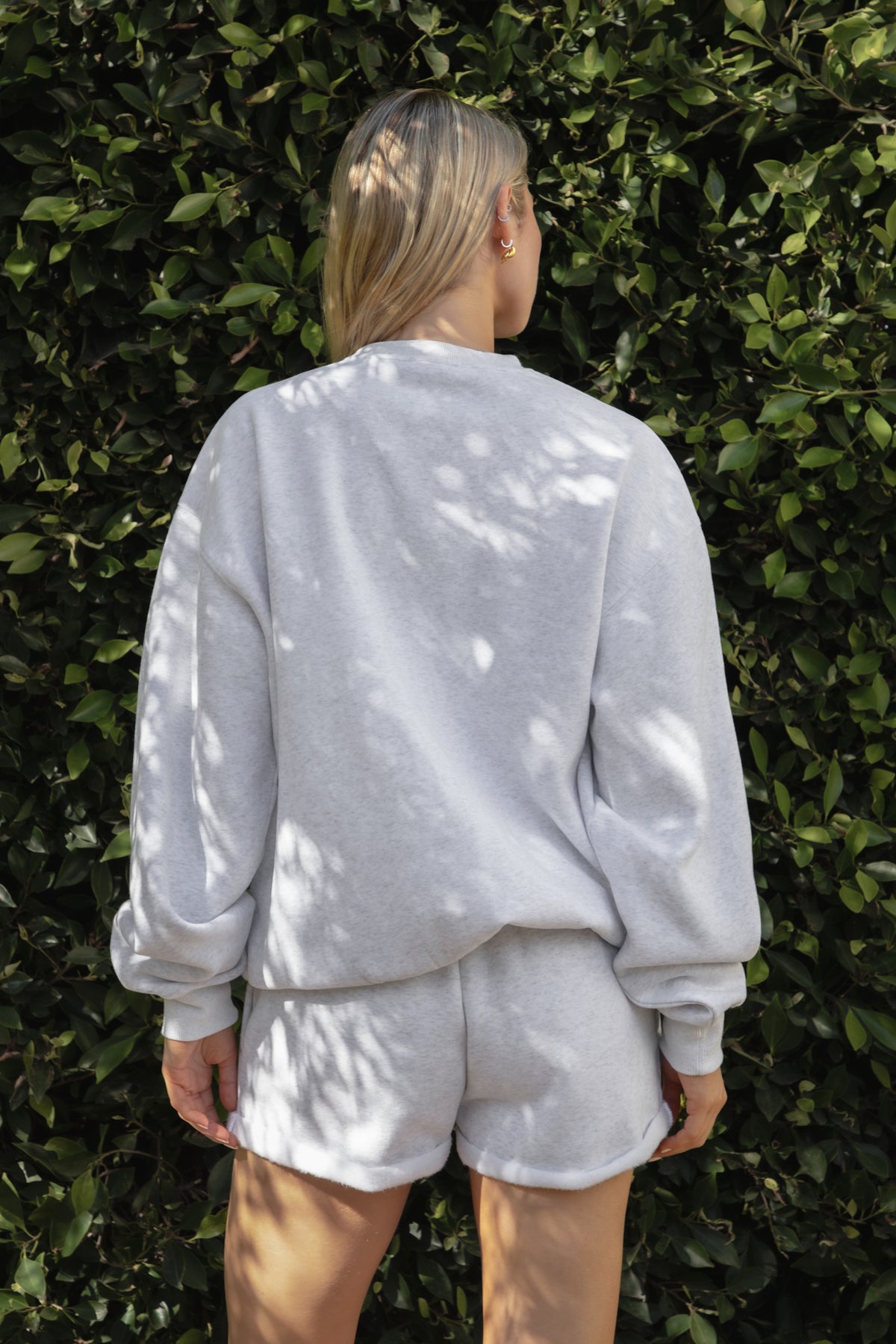 Woman wearing the TIC TOC TJ31656 LANI ash cloud fleece crew neck pullover sweatshirt featuring dropped shoulders, ribbed trim, and cozy fleece fabric with the matching bottoms against a green leafy background.