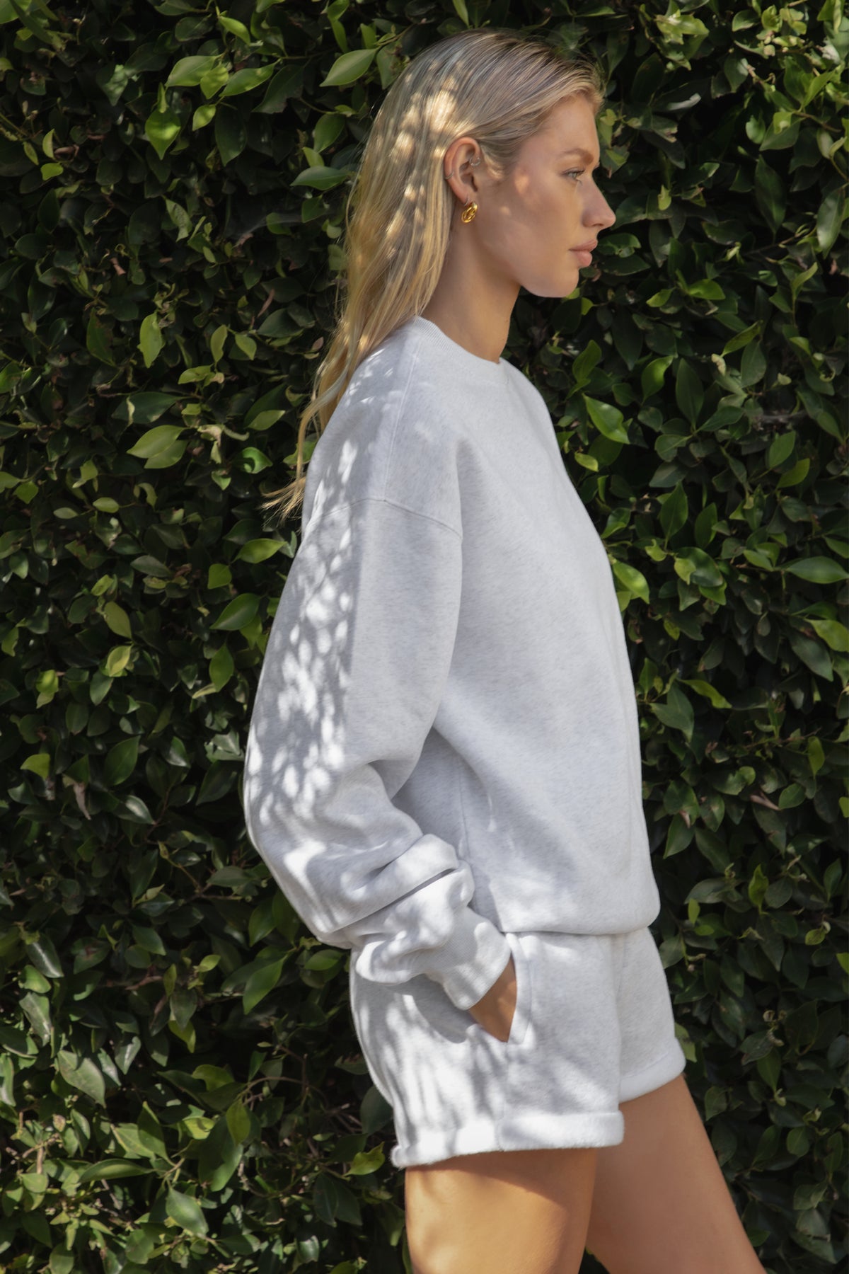 Woman wearing the TIC TOC TJ31656 LANI ash cloud fleece crew neck pullover sweatshirt featuring dropped shoulders, ribbed trim, and cozy fleece fabric with the matching bottoms against a green leafy background.