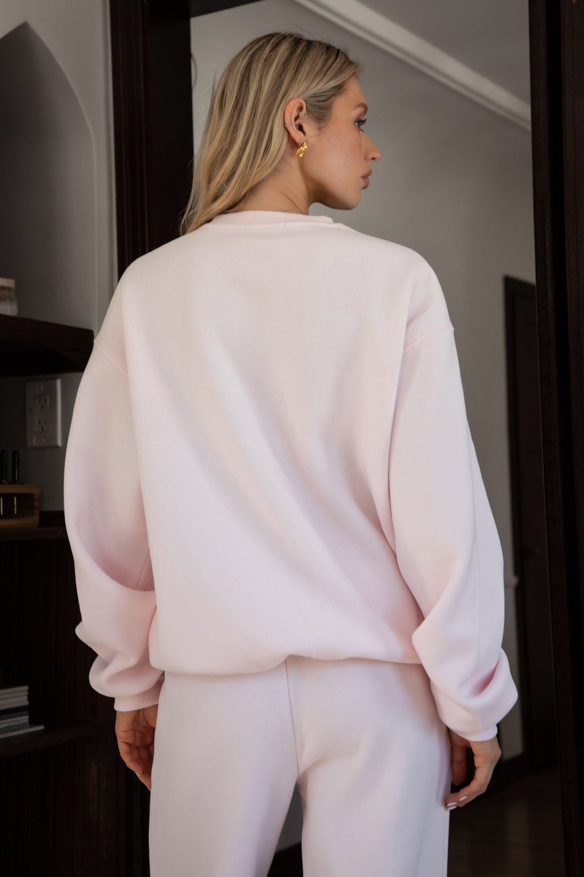 Woman wearing the TIC TOC TJ31656 LANI pale pink cloud fleece crew neck pullover sweatshirt featuring dropped shoulders, ribbed trim, and cozy fleece fabric with the matching bottoms indoors.