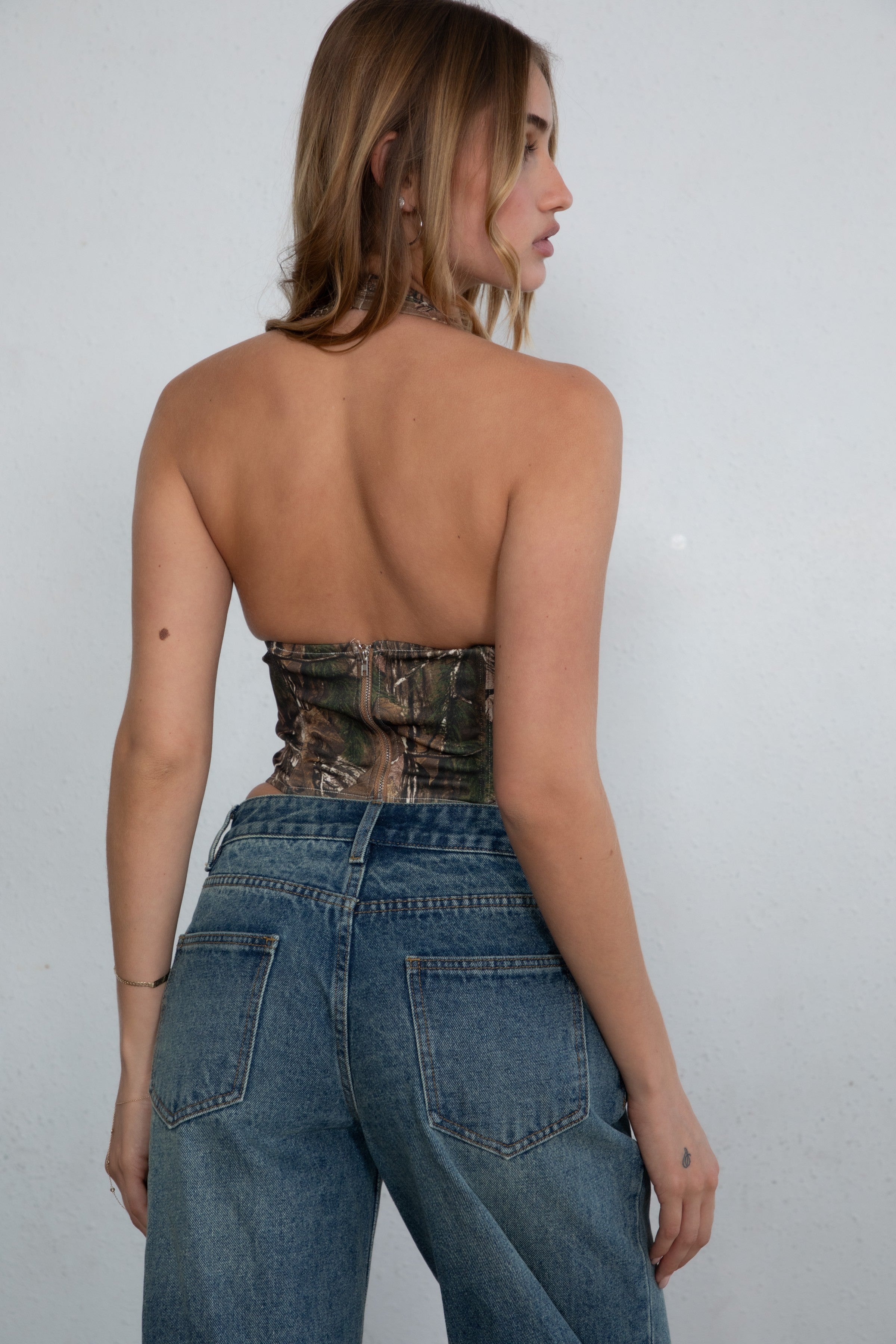 Woman standing in a wooden cabin doorway wearing TIC TOC CAMOU Camo Print Halter Corset Top in brown with exposed chest, curved hem, and stretch fit paired with denim shorts, and cowboy boots