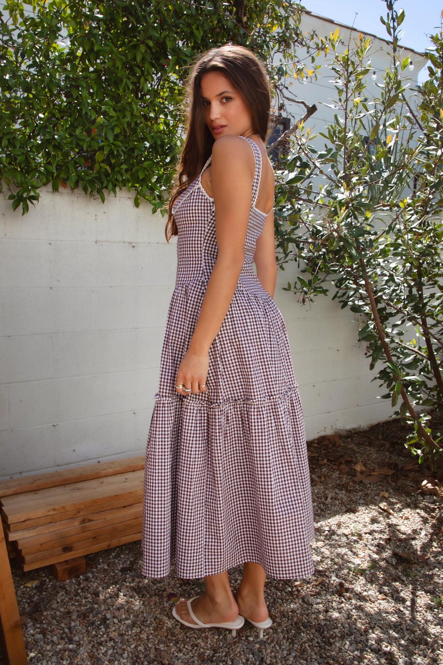A woman standing outdoors wearing a purple gingham A-line maxi dress with a square neckline, fixed shoulder straps, contrast lace trim, a Peter Pan collar with a satin bow at the center, and a hidden zipper closure at the side. The dress has a shaped waist and a V-cut hip line, made of double-lined flowy fabric.