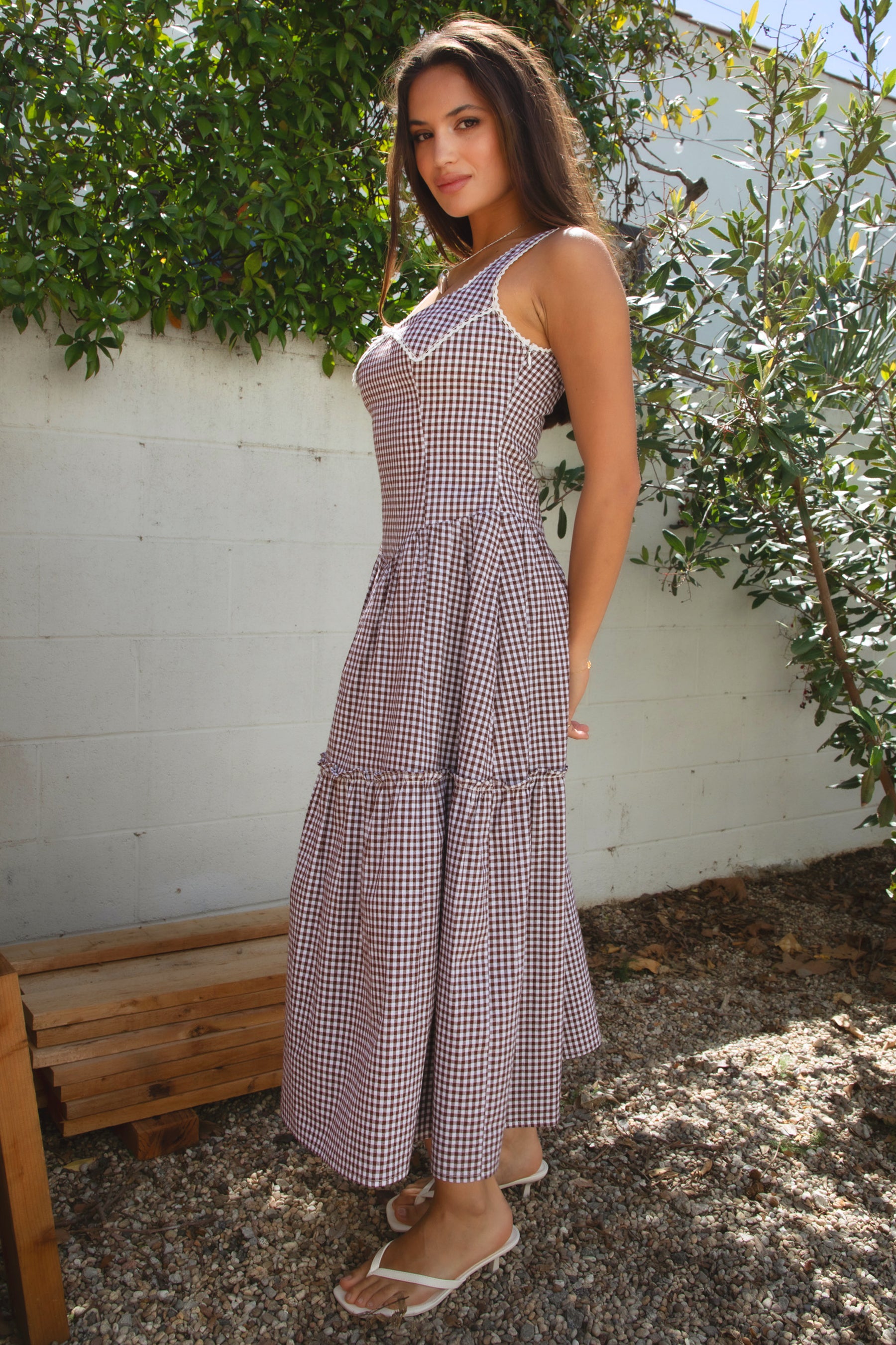 A woman standing outdoors wearing a purple gingham A-line maxi dress with a square neckline, fixed shoulder straps, contrast lace trim, a Peter Pan collar with a satin bow at the center, and a hidden zipper closure at the side. The dress has a shaped waist and a V-cut hip line, made of double-lined flowy fabric.
