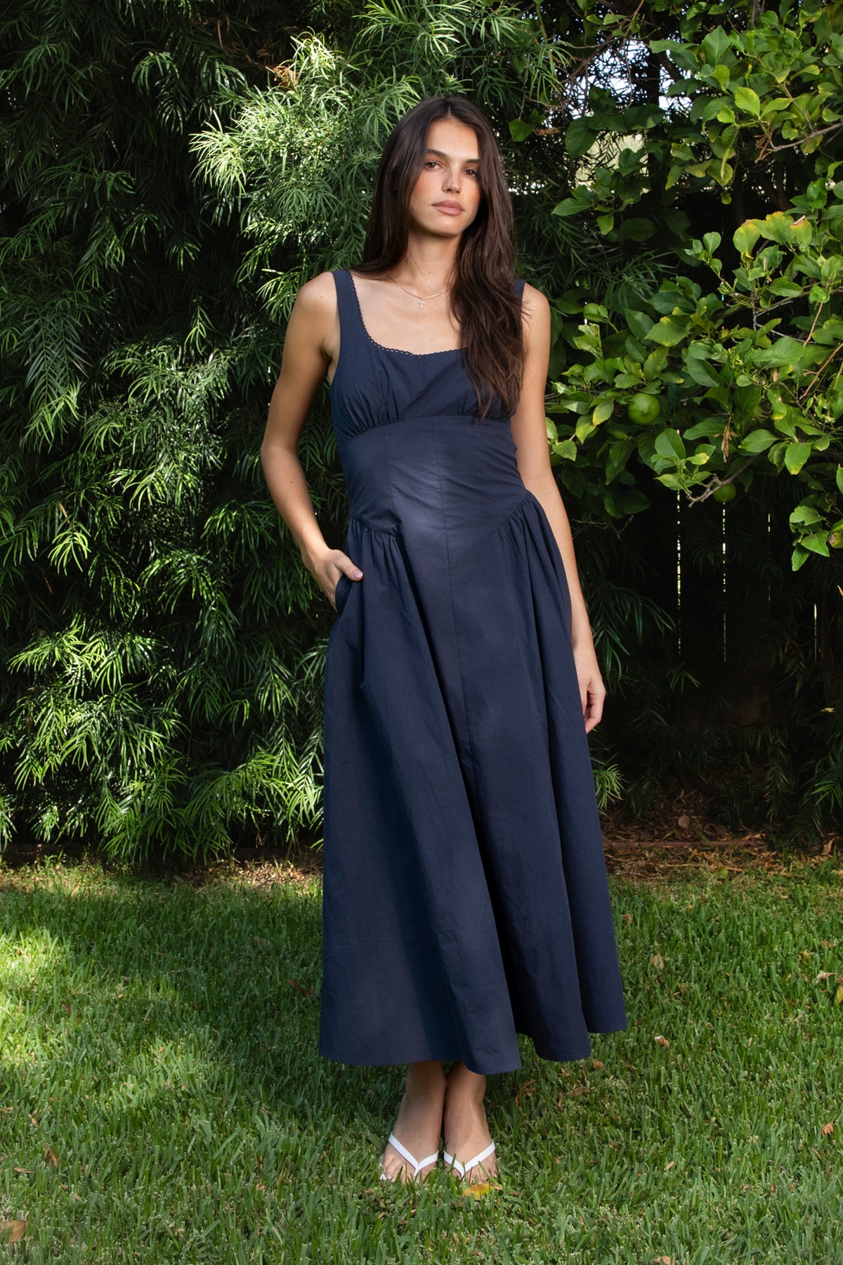 Woman wearing NAVYA scoop neck cotton poplin midi dress featuring soft smooth cotton poplin fabric, feminine scoop neckline with delicate picot trim, wide shoulder straps with adjustable back detail, gathered bust with built in bra cups, fitted waistline with shaped seam detailing, smocked back panel, and full flared midi length skirt with soft side gathers and rayon lining.