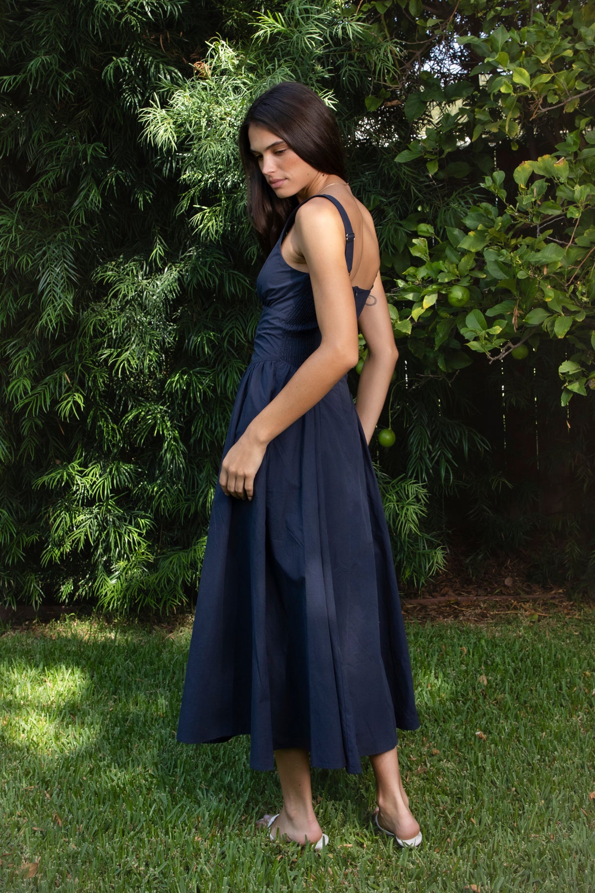 Woman wearing NAVYA scoop neck cotton poplin midi dress featuring soft smooth cotton poplin fabric, feminine scoop neckline with delicate picot trim, wide shoulder straps with adjustable back detail, gathered bust with built in bra cups, fitted waistline with shaped seam detailing, smocked back panel, and full flared midi length skirt with soft side gathers and rayon lining.