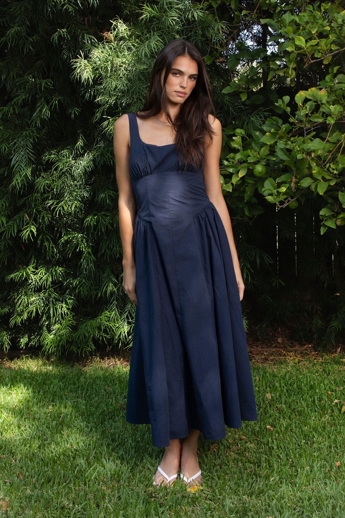 Woman wearing NAVYA scoop neck cotton poplin midi dress featuring soft smooth cotton poplin fabric, feminine scoop neckline with delicate picot trim, wide shoulder straps with adjustable back detail, gathered bust with built in bra cups, fitted waistline with shaped seam detailing, smocked back panel, and full flared midi length skirt with soft side gathers and rayon lining.
