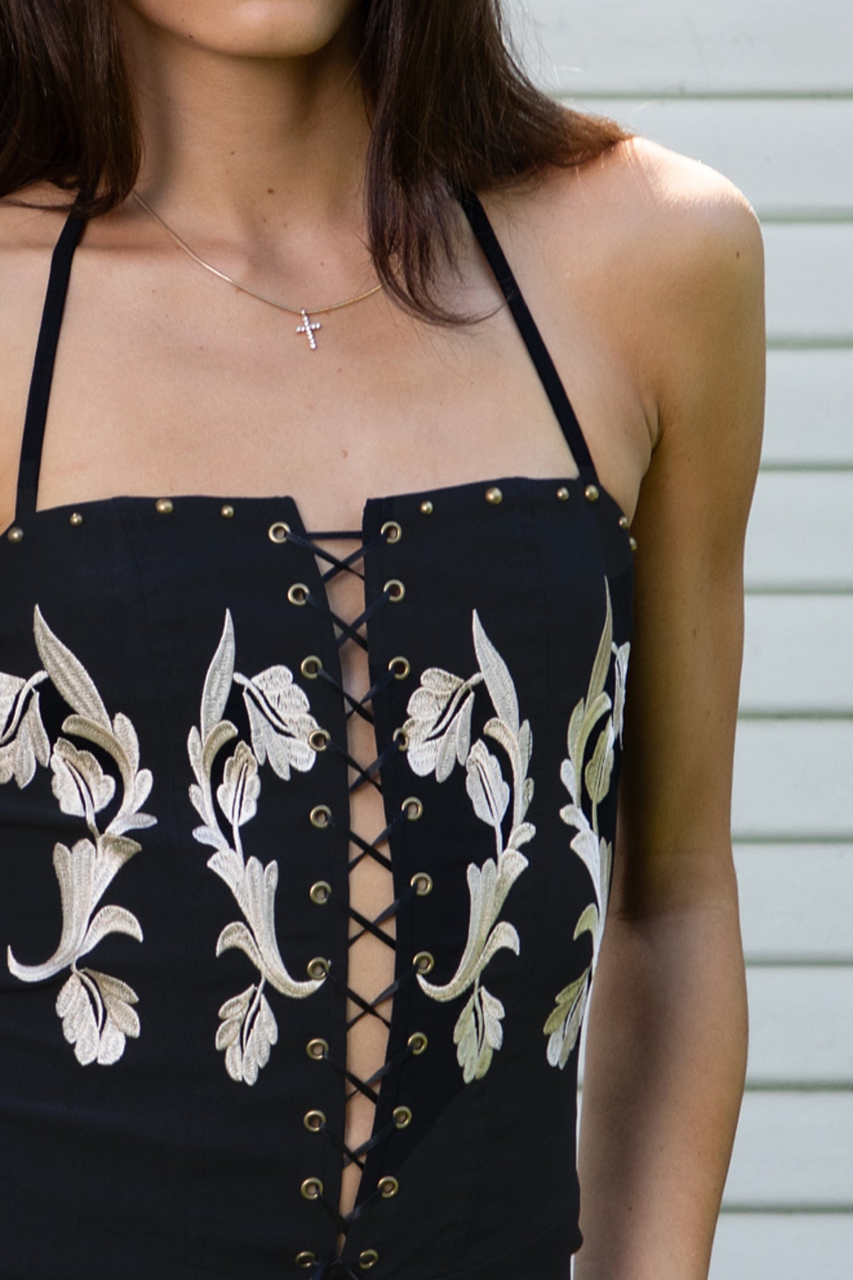 Woman wearing CLEMENTINE embroidered lace up halter mini dress featuring structured stretch fabric, fitted bodycon silhouette, straight neckline with adjustable halter tie straps, floral embroidery with western inspired details, deep center front lace up design with metal eyelets, gold tone stud embellishments along the neckline and hem, corset inspired contoured seaming, exposed back, and short mini length.