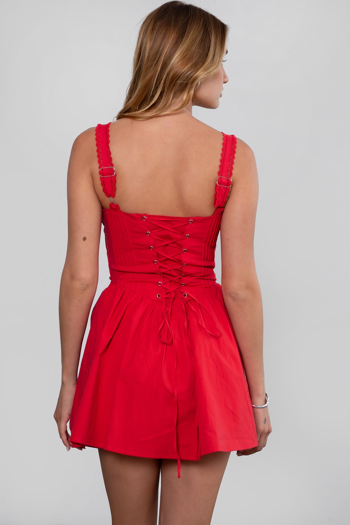 A woman standing in a studio setting, wearing a red mini dress with adjustable lace shoulder straps, a cinched bust with bow detail, and a short A-line skirt. The dress features a corset-style bodice and lace overlay at the waist.