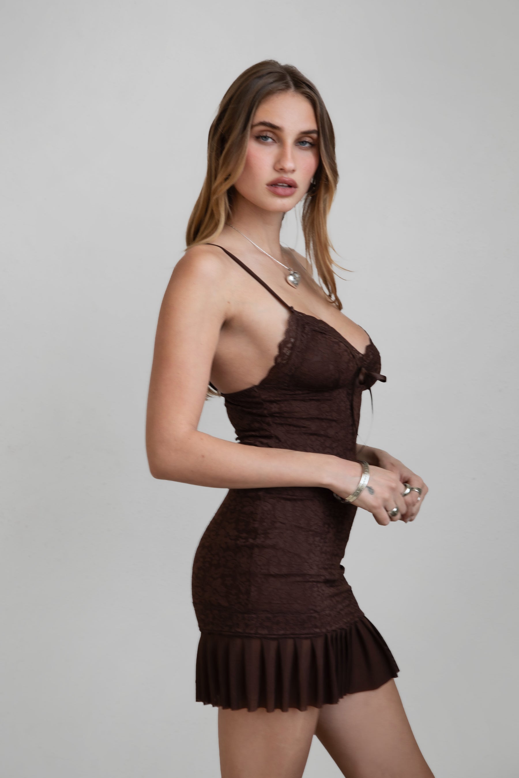 Woman in TIC TOC CARLITA Lace Bodycon Mini Dress in brown featuring a deep V neckline, satin bow detail, and pleated mesh hem in a plain background setting