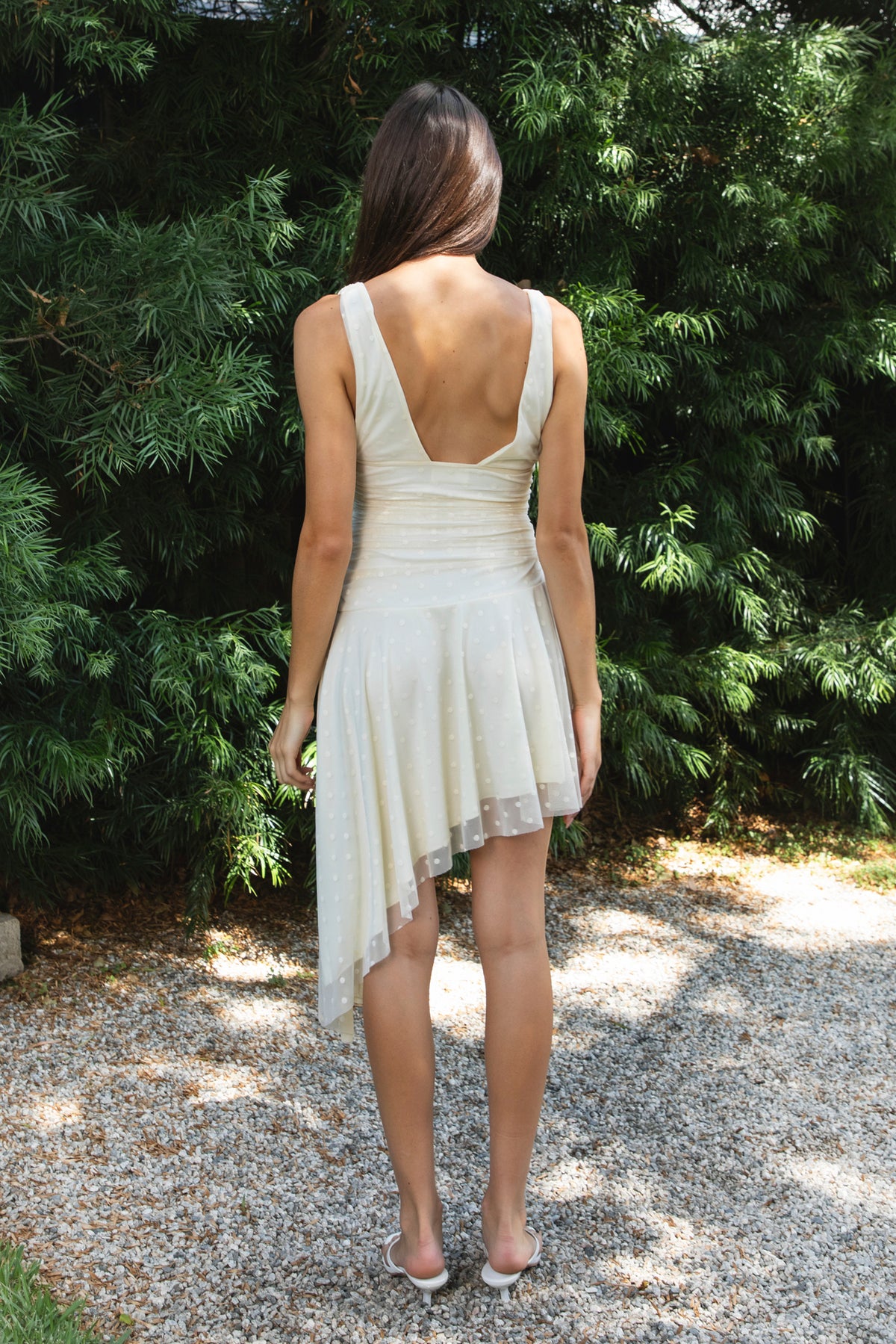 Woman wearing FIORA flocked dot asymmetrical mini dress featuring soft flocked dot mesh, sleeveless silhouette, deep V neckline with gathered bust detail, ruched fitted waist, low V back neckline, asymmetrical handkerchief hem with extended side drape, and soft interlock jersey lining.